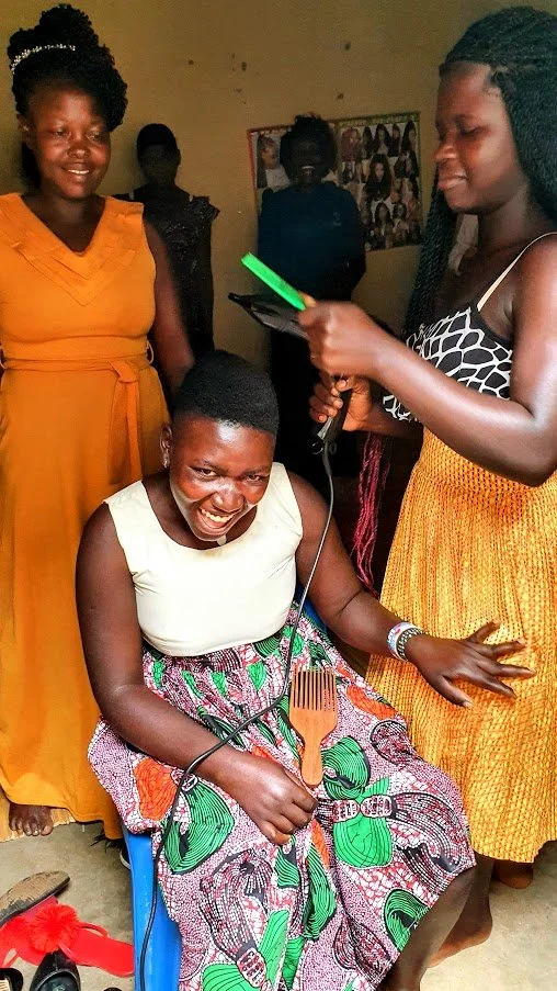 A joyful woman sitting in a wheelchair, holding a hairbrush, surrounded by four women at a small group of trainee hairdressers near Tororo all smiling and dressed in colorful dresses.
