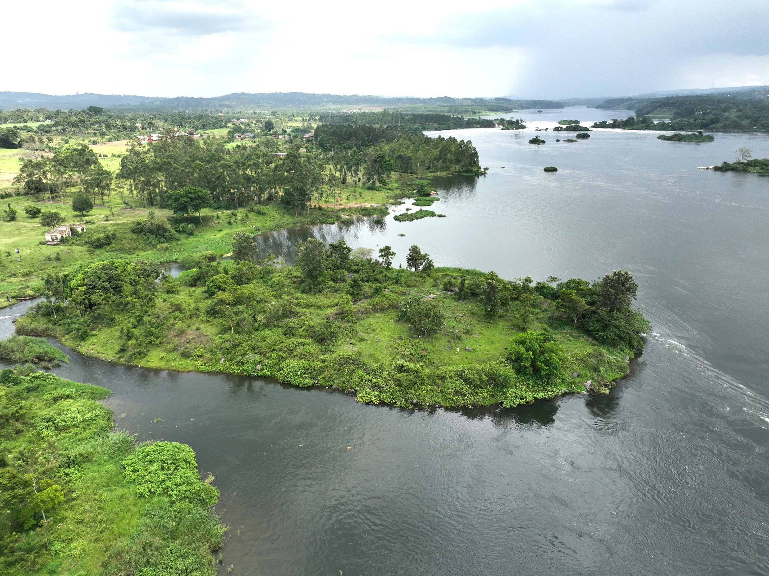 A wide river with small islands and lush green trees along its banks, with distant hills and a cloudy sky in the background.
