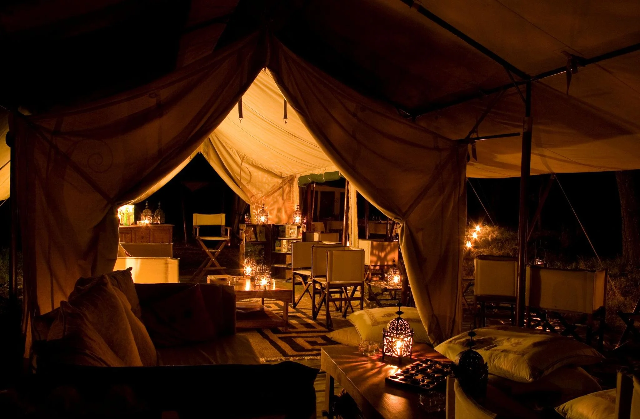 Nighttime scene inside a tent with warm lighting, candle lanterns, armchairs, and decorative lamps creating a cozy ambiance.