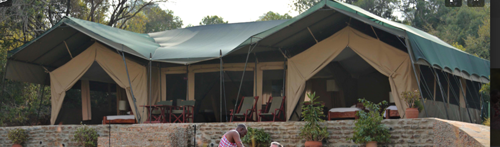 A large canvas tent on a raised brick platform with outdoor furniture and potted plants, set in a wooded area.