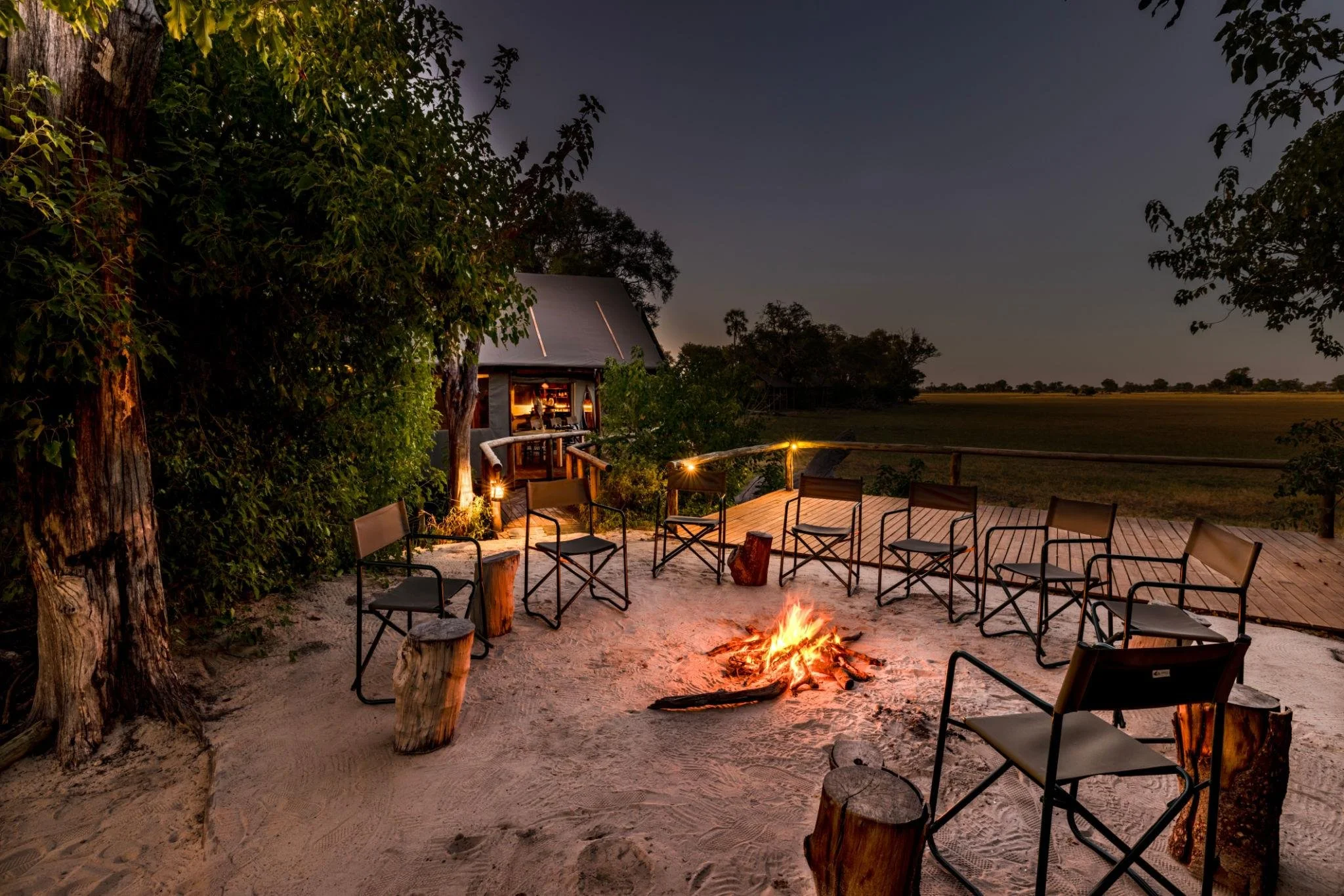 An outdoor seating area with chairs and a campfire on a sandy surface at night. There are trees on the left and right, a small structure with a lit interior in the background, and string lights illuminating the area.