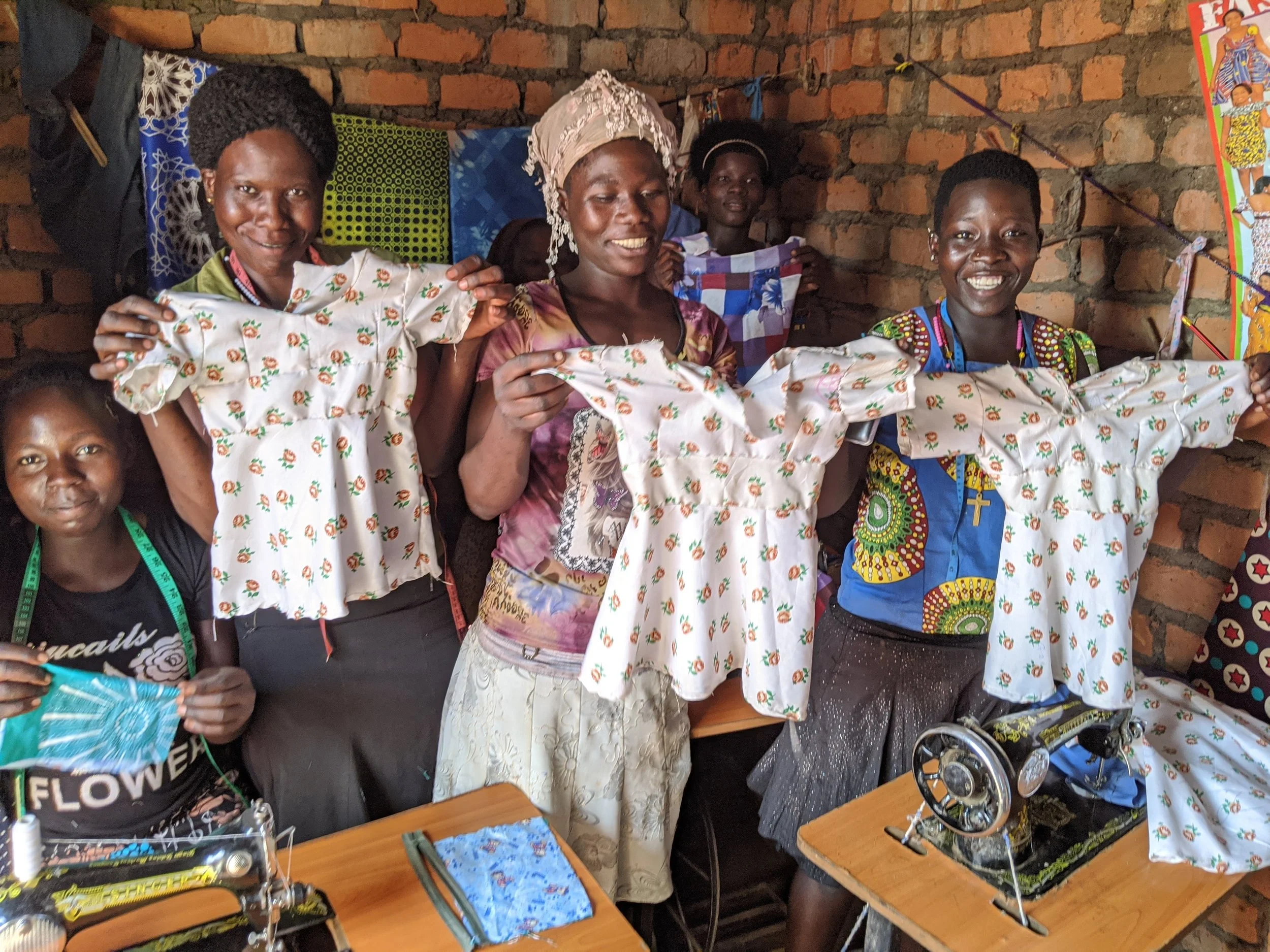 Women and girls in colorful traditional clothing holding embroidered fabrics in a brick-walled room, with tools and equipment on the table.
