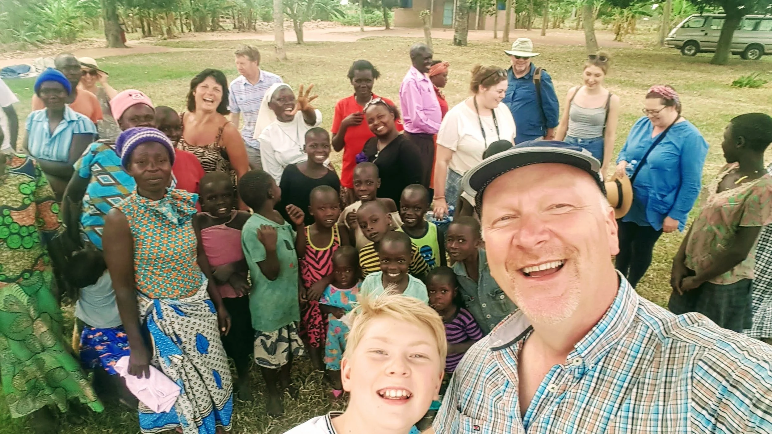 A group of diverse children and adults smiling and posing for a selfie outdoors in a grassy area with trees, vehicles, and a building in the background.
