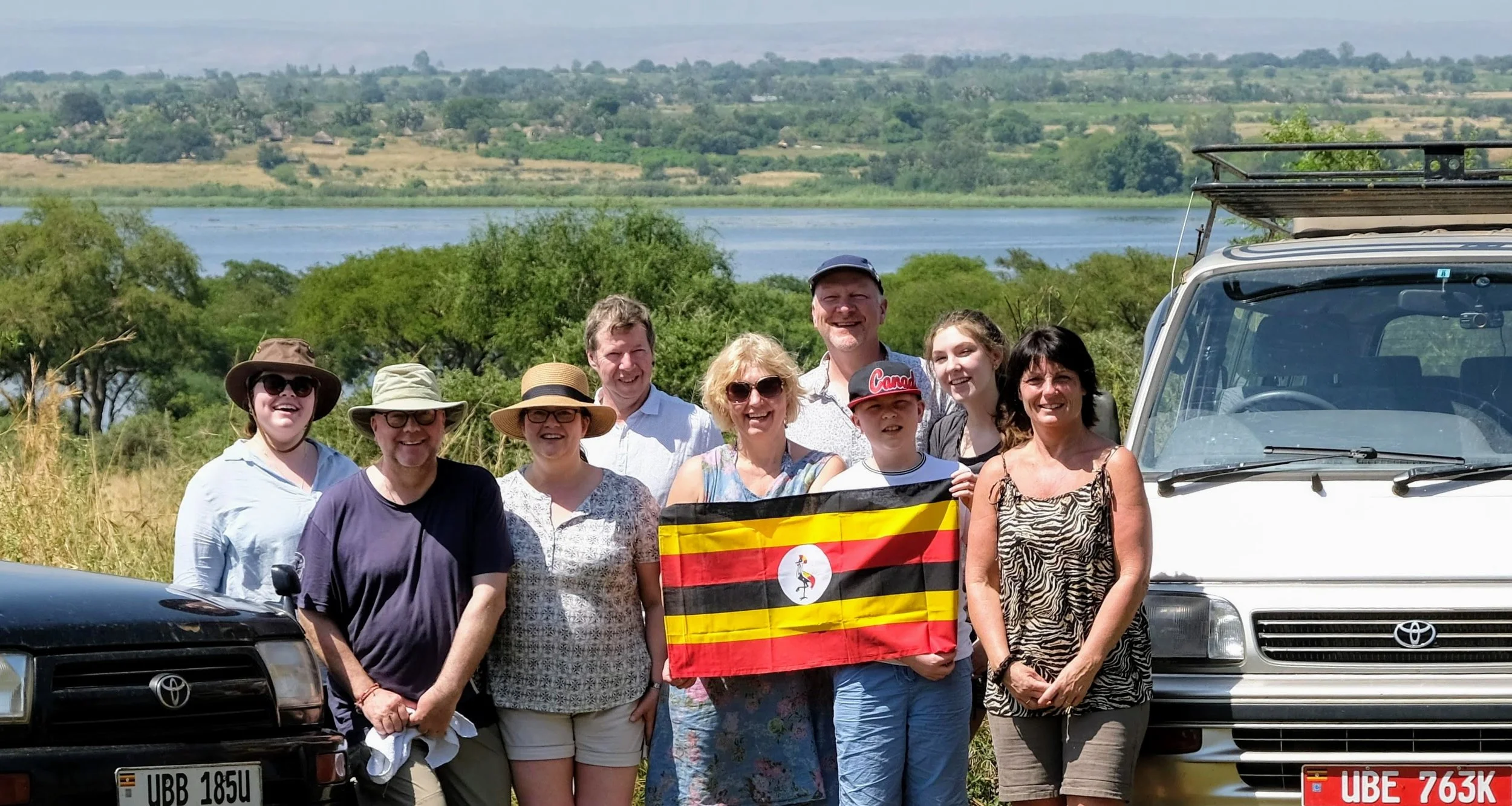 A group of eleven people, including children and adults, standing outdoors, holding a Ugandan flag, with cars and a river in the background on a sunny day.
