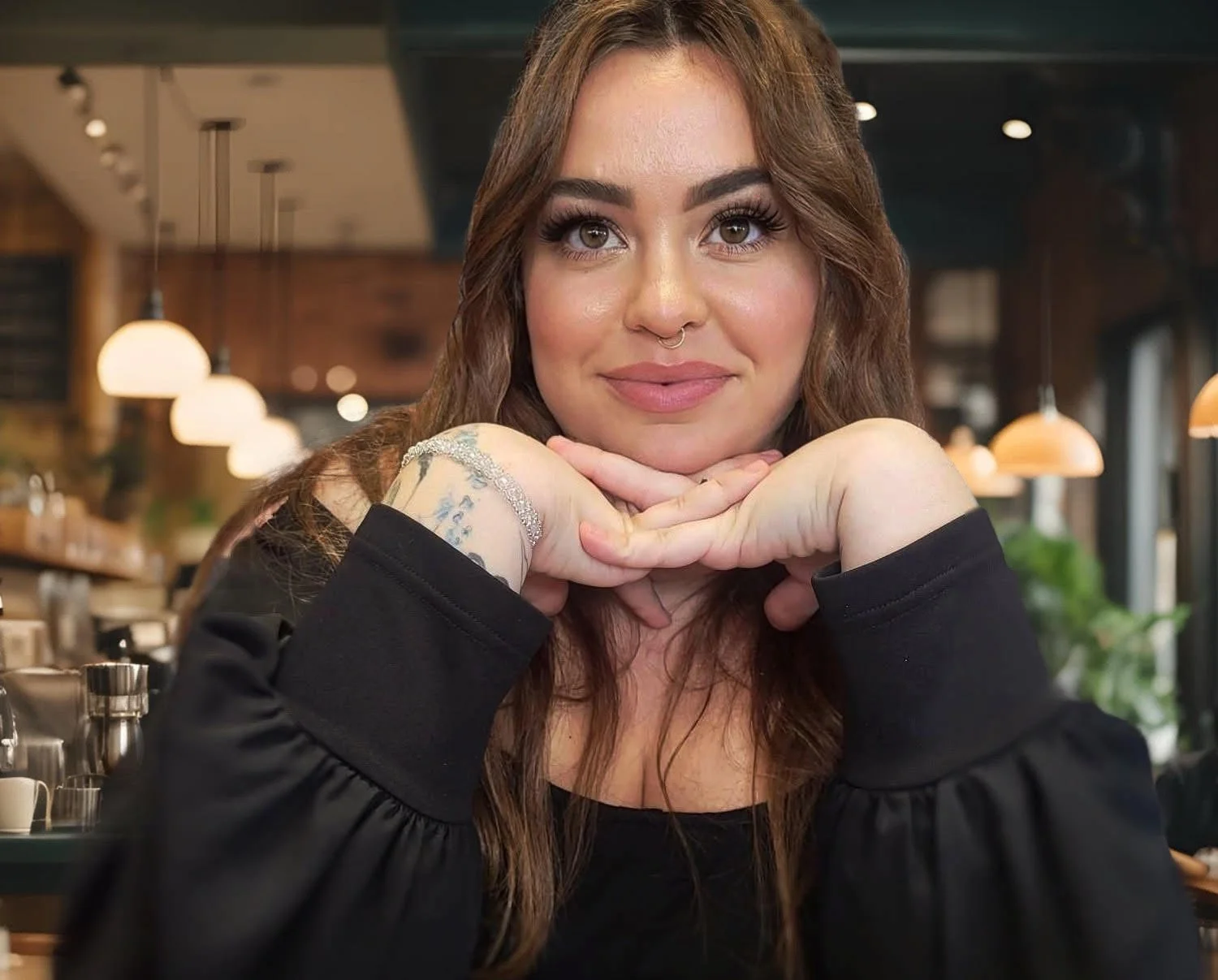 A woman with long brown hair, brown eyes, and makeup, resting her chin on her hands and smiling, seated in a cafe or restaurant with hanging lights and a cozy ambiance.