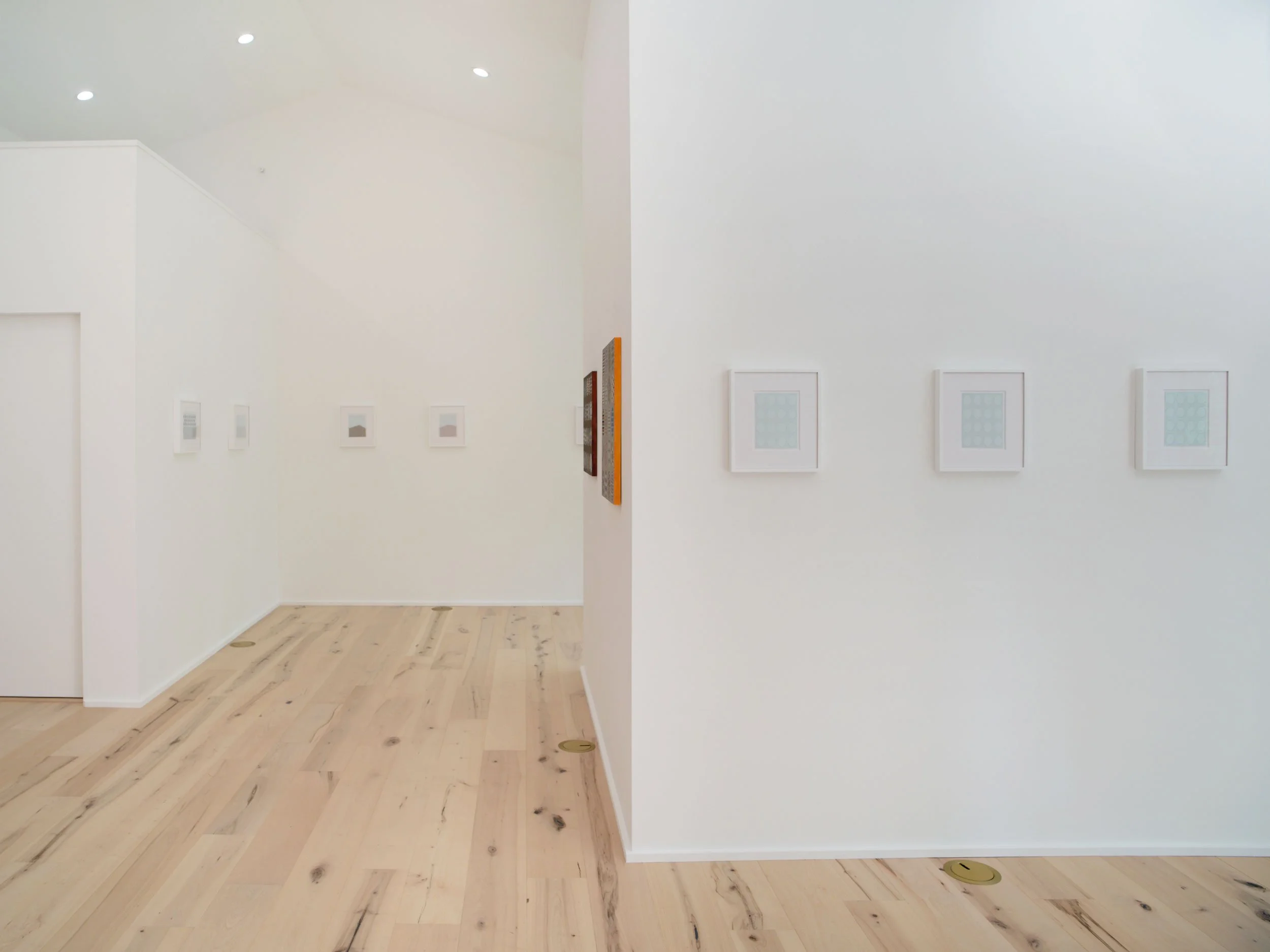 A minimalist art gallery with white walls displaying several pieces of framed artwork arranged in a row. The gallery features a light wooden floor and bright ceiling lighting.