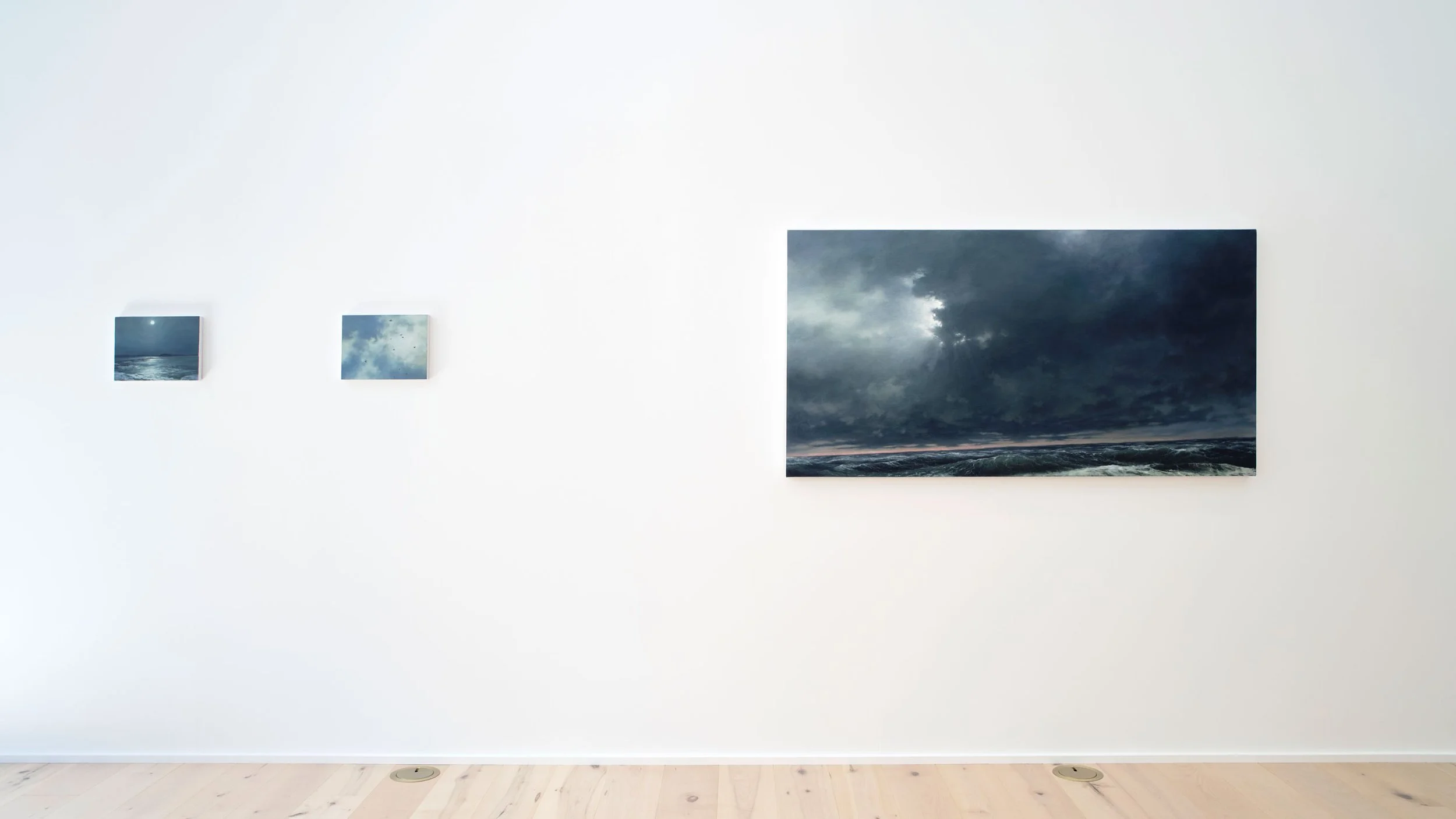 An art gallery with white walls displaying landscape photographs, natural light coming through a large window, and wooden flooring.
