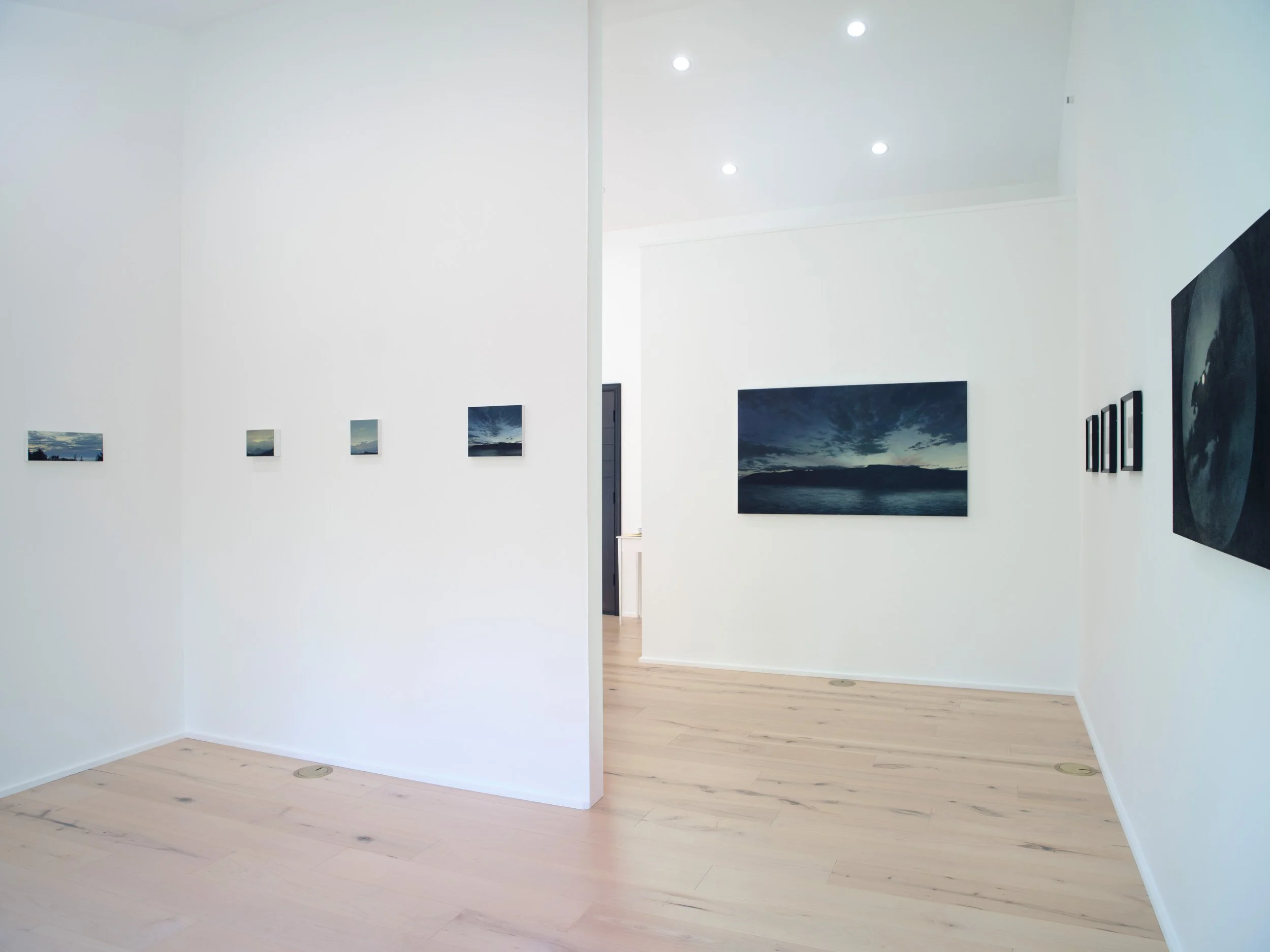 A modern art gallery with white walls, wooden flooring, and wall-mounted landscape photographs. Some photograph frames are visible, and the room is well-lit with recessed lighting in the ceiling.