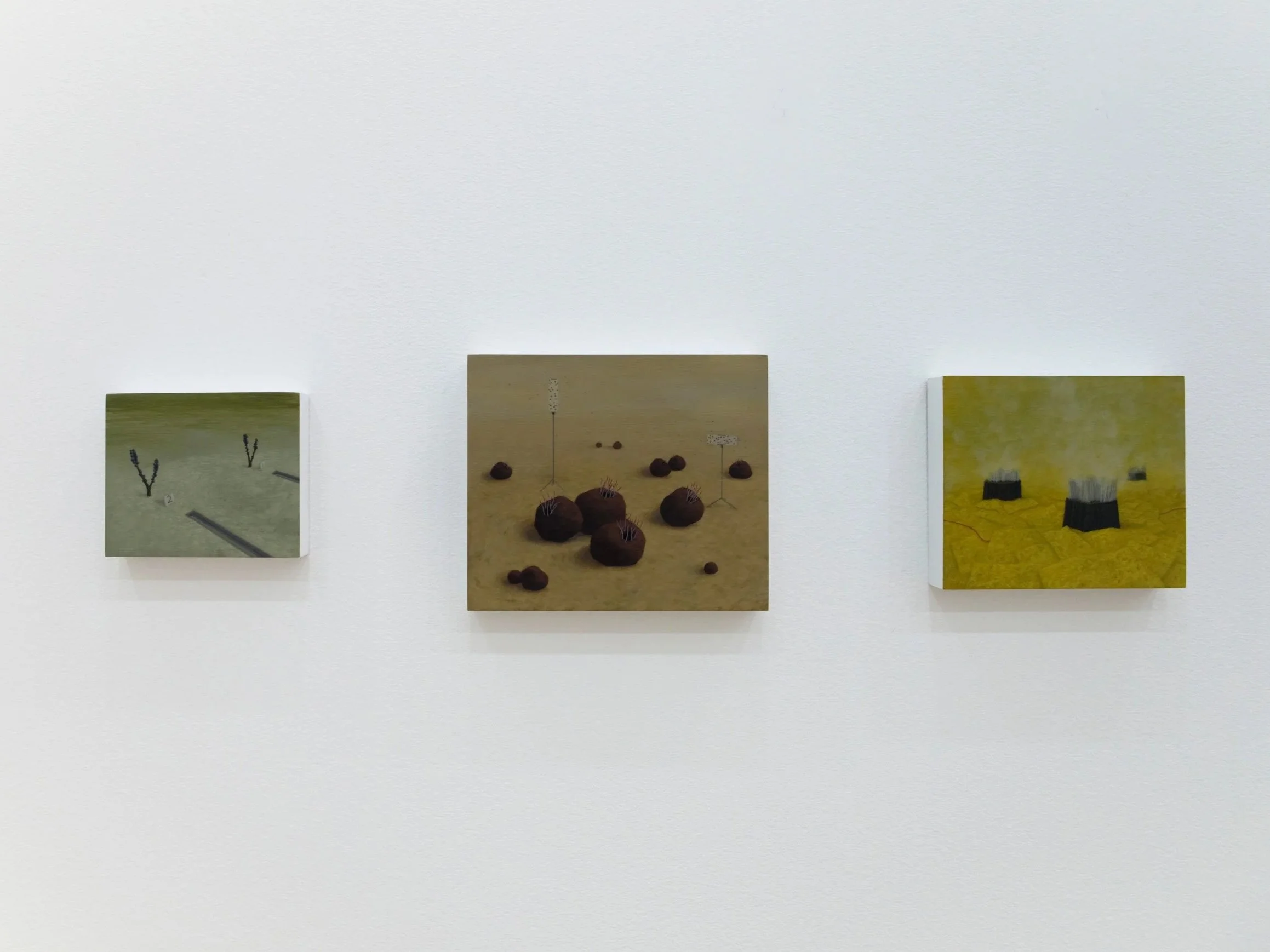Three small art pieces displayed on a white gallery wall, each featuring abstract or minimalistic designs with muted colors.