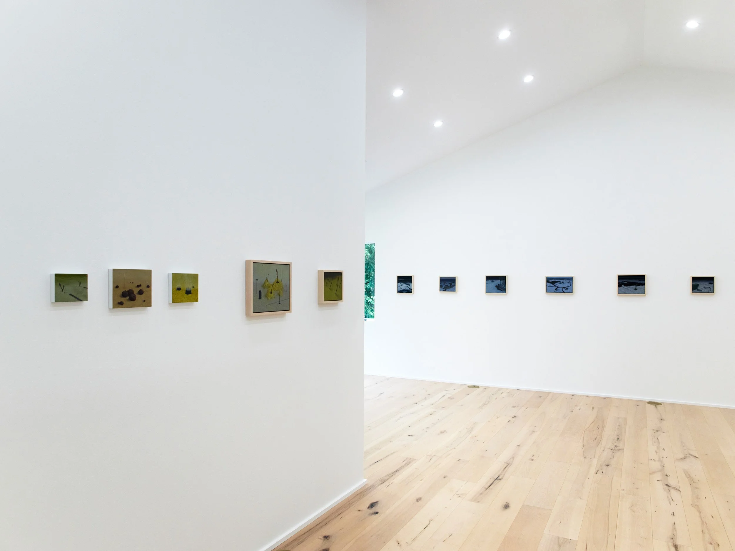 An art gallery with white walls displaying framed pictures. The left section showcases a few small artworks with greenery outside visible through a small window. The right section features a row of small framed pieces on a plain white wall.