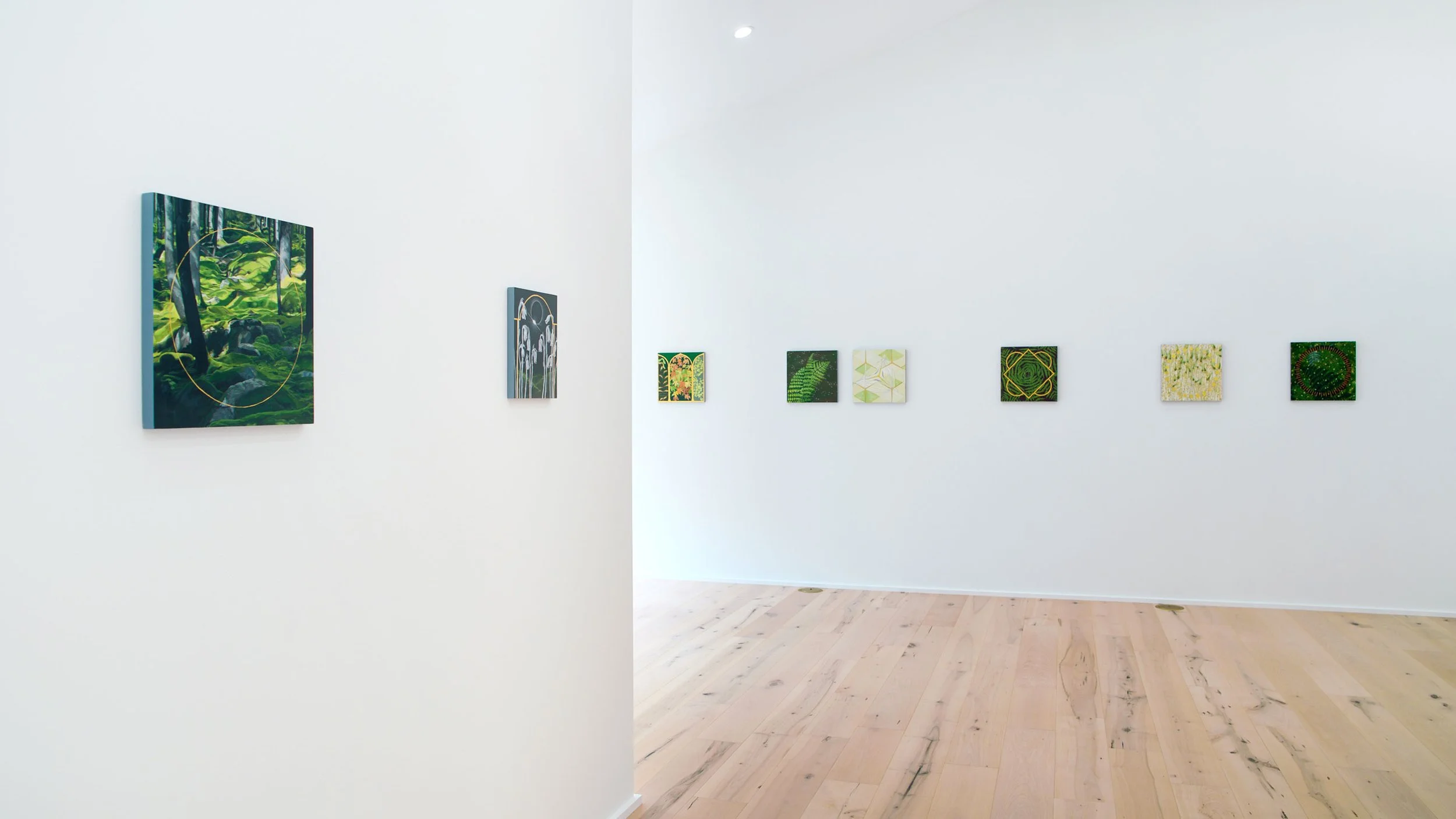A white-walled art gallery with displayed paintings, showing a forest scene, modern abstract designs with greenery, yellow, and black colors, and a circular pattern, with a wooden floor.