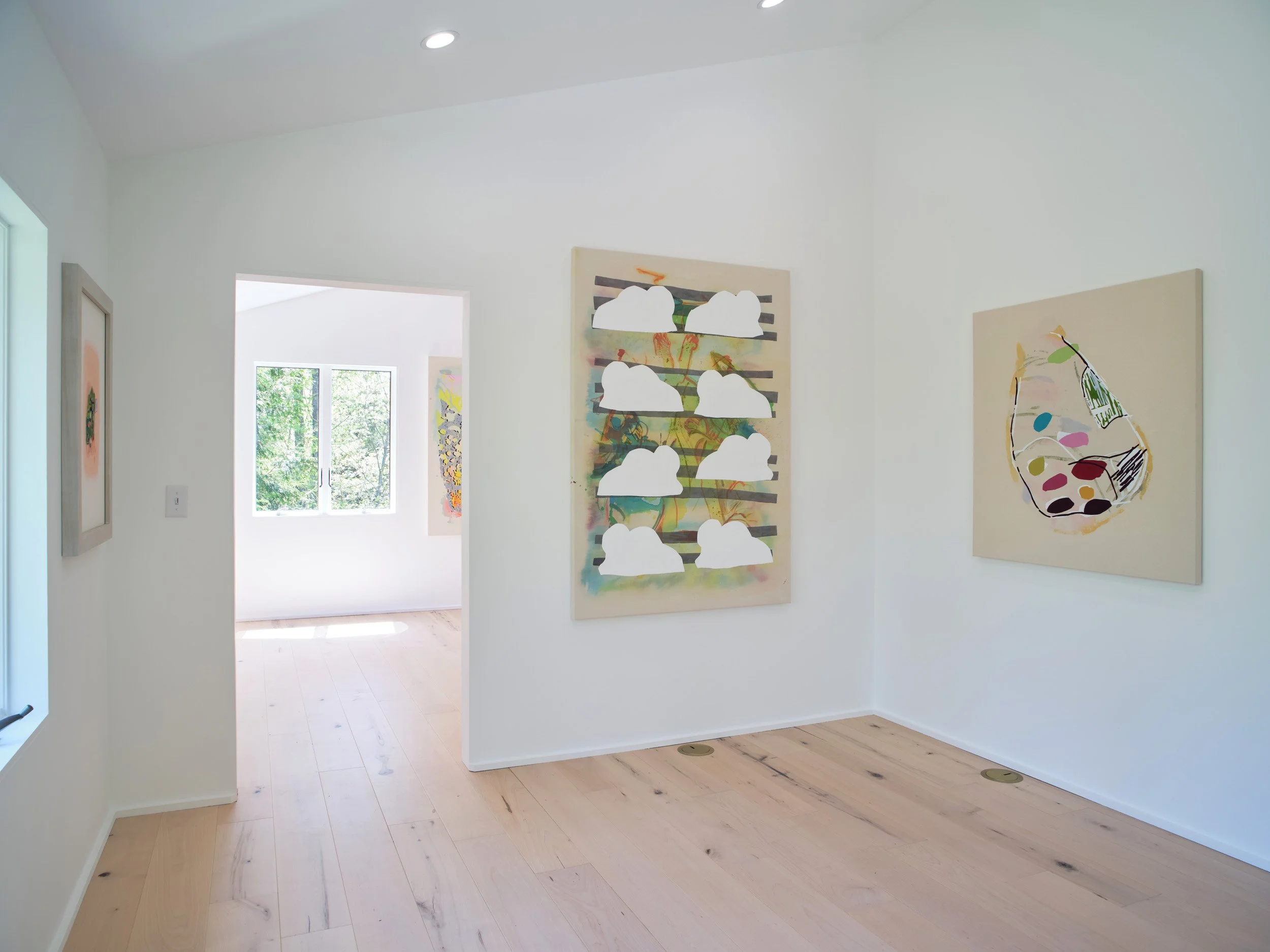 Inside view of an art gallery with white walls, wooden floors, and colorful abstract paintings displayed. Light from windows highlights the artwork and a doorway leads to a bright room with more artwork.