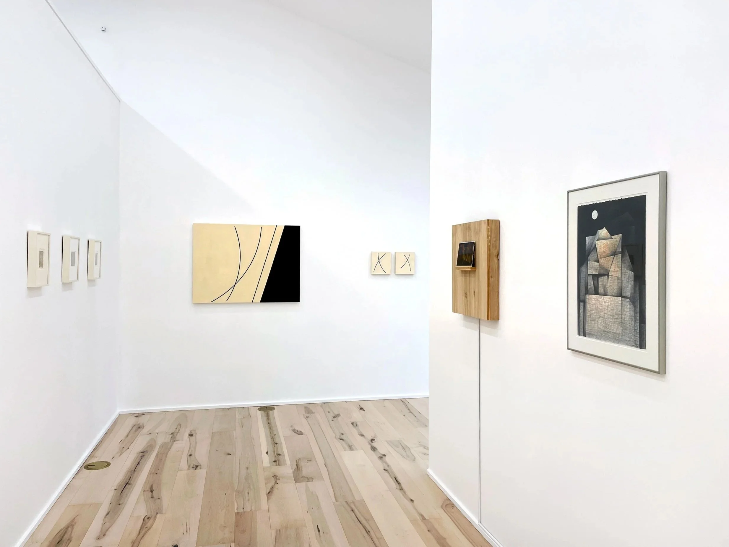 An art gallery with white walls displaying various artworks including framed paintings and abstract art, with light wood flooring and a mirror reflecting part of the exhibit.