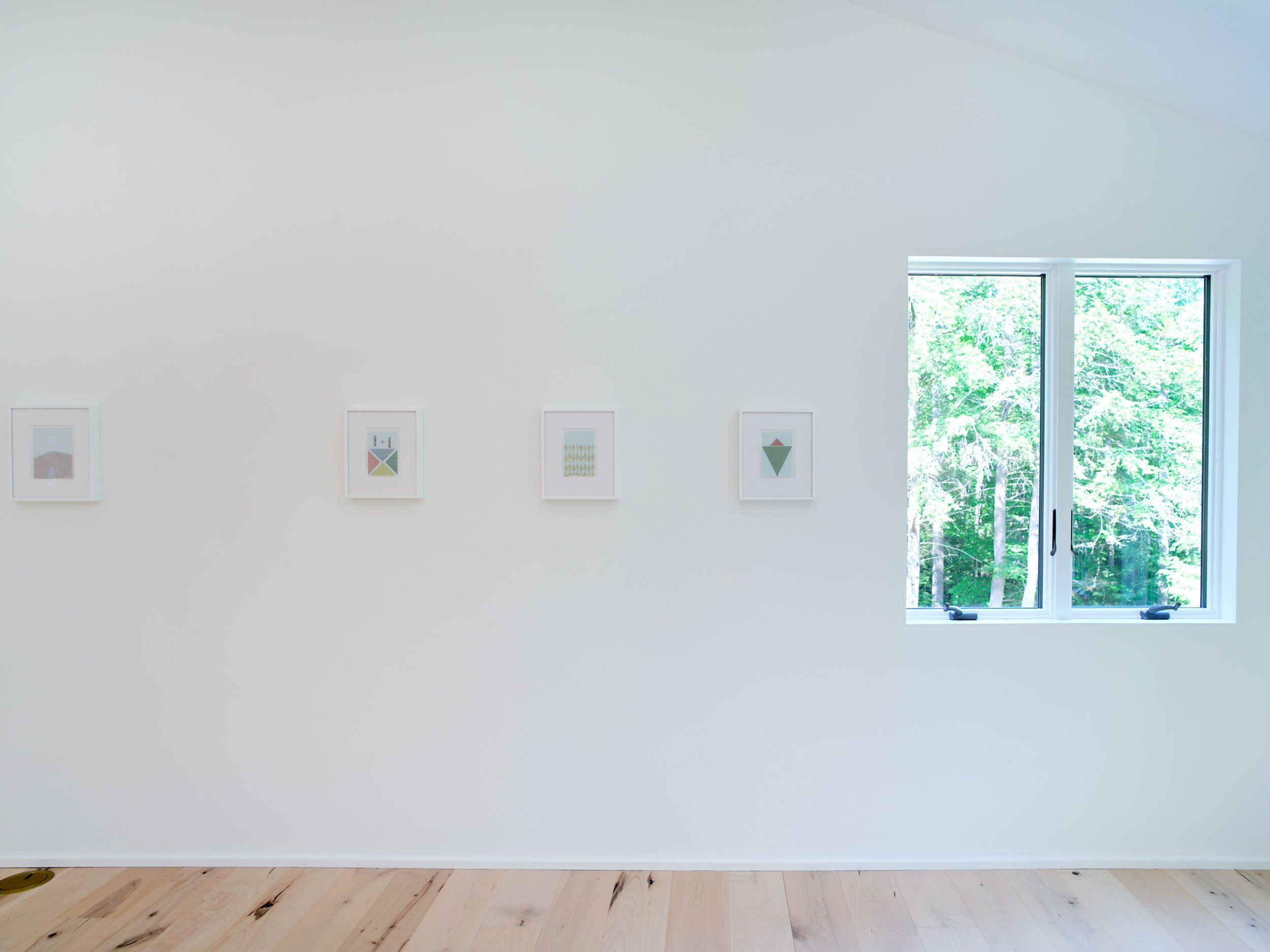 A plain white wall with four minimalist framed artworks and a large window showing green trees outside.