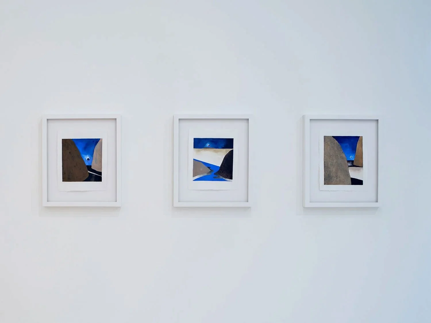 Three framed landscape paintings hung on a white gallery wall, featuring rocky cliffs, blue skies, and a river flowing between the rocks.