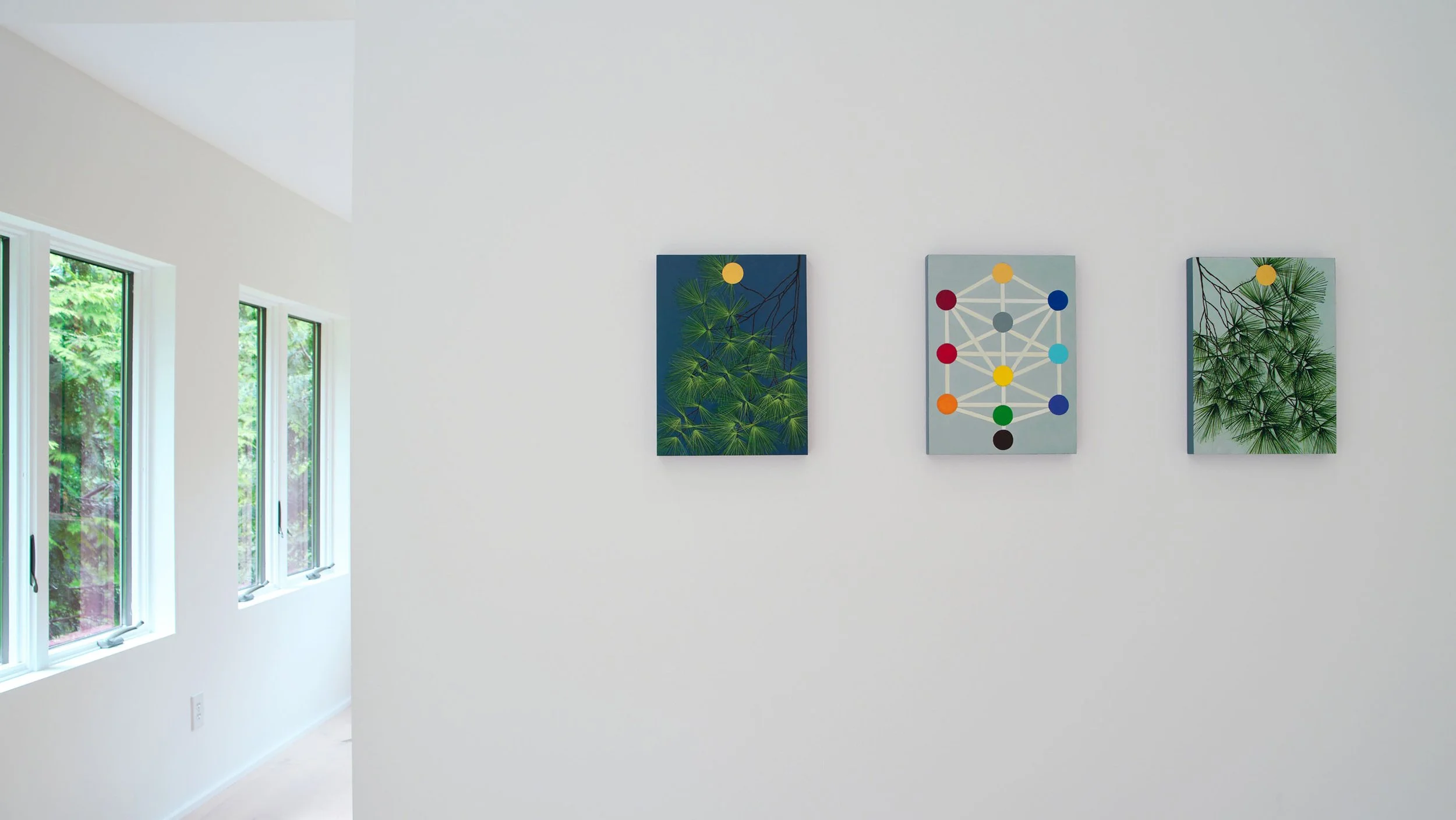 Three botanical-themed art pieces hanging on a white wall near large windows showing outdoor greenery.