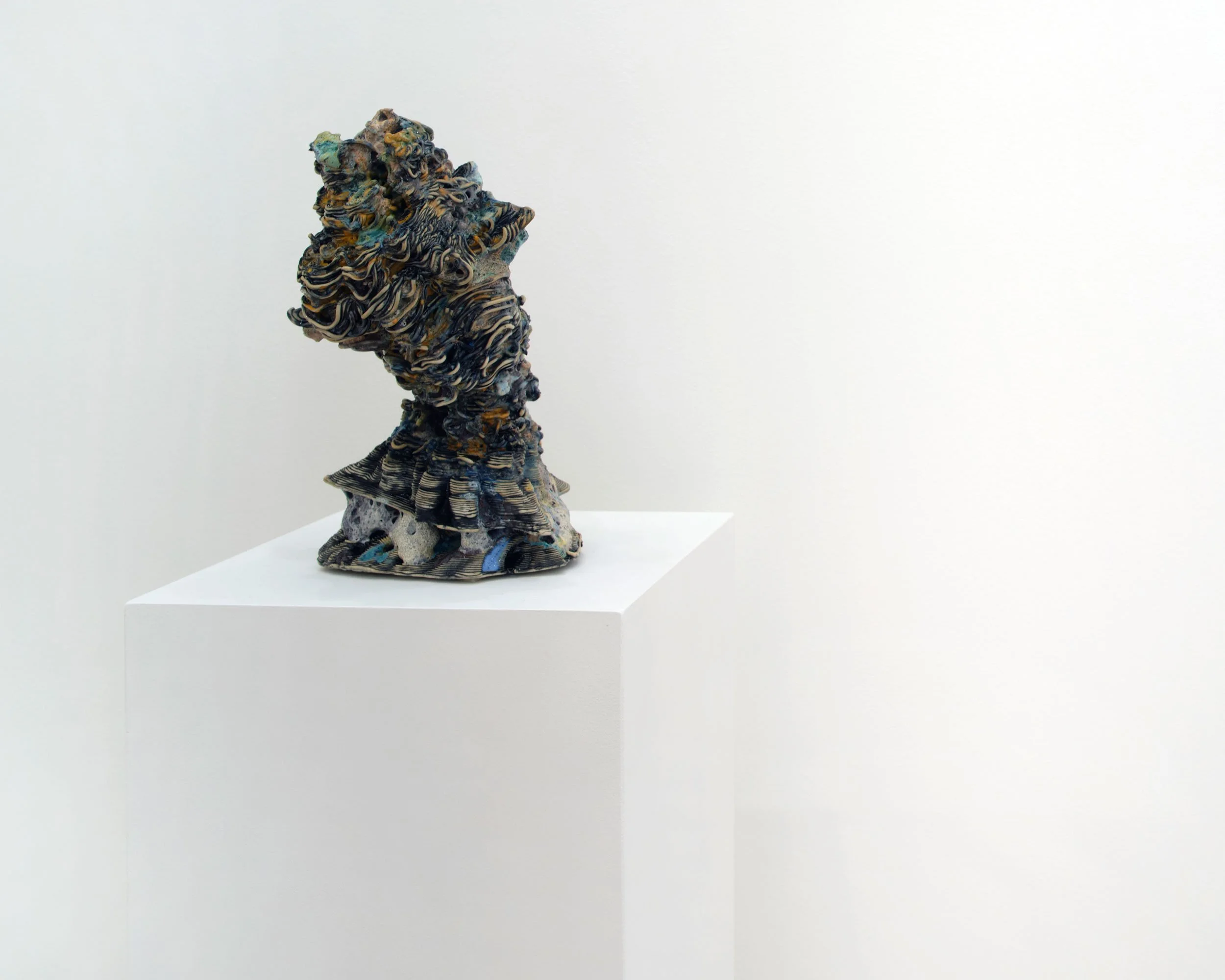 A mixed media sculpture resembling a bust of a person, made with various textured and colorful materials, displayed on a white pedestal against a plain white wall.