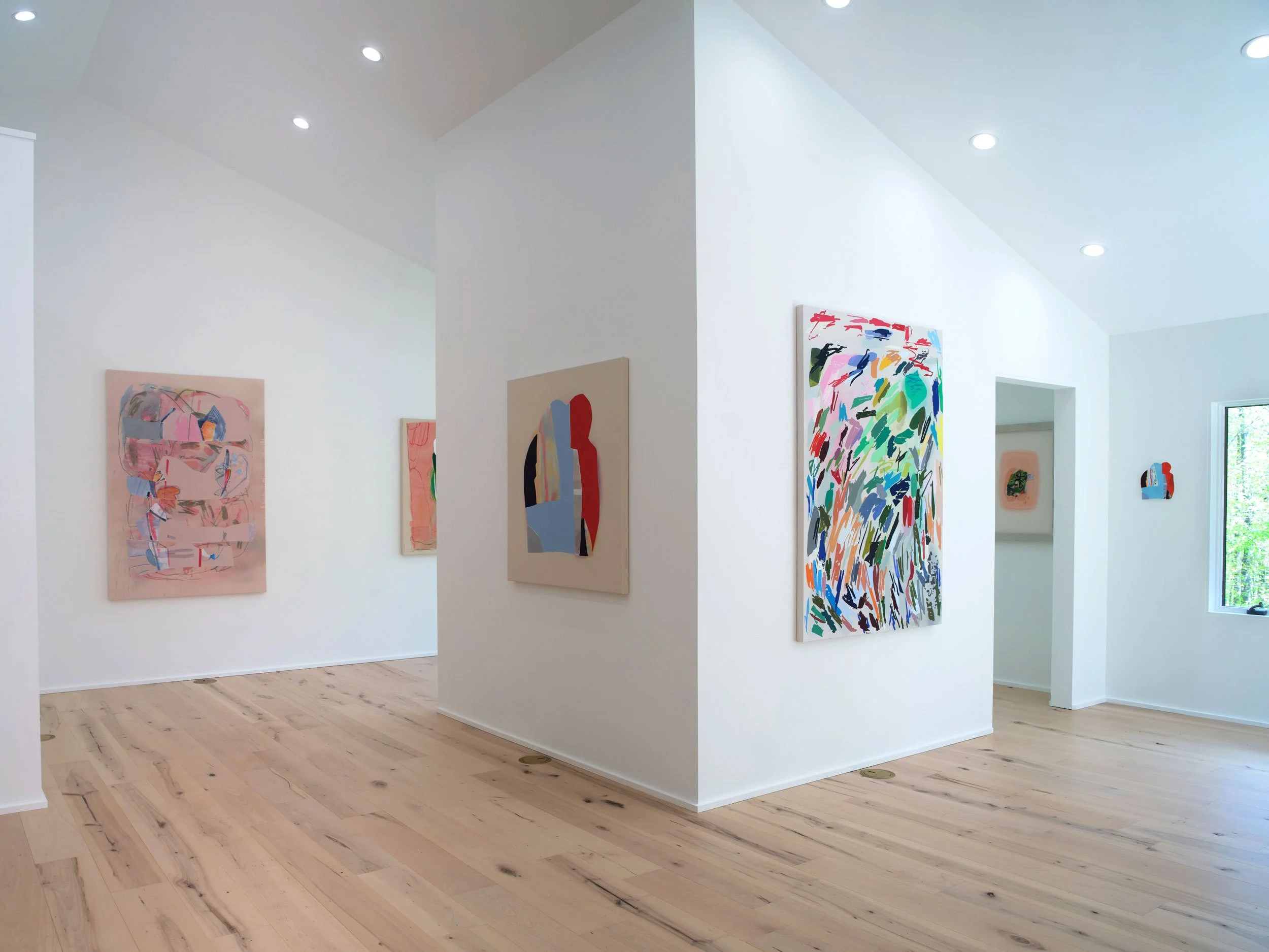 An art gallery with white walls and light wooden flooring, displaying colorful abstract paintings, illuminated by ceiling lights, with a window visible on the right side.