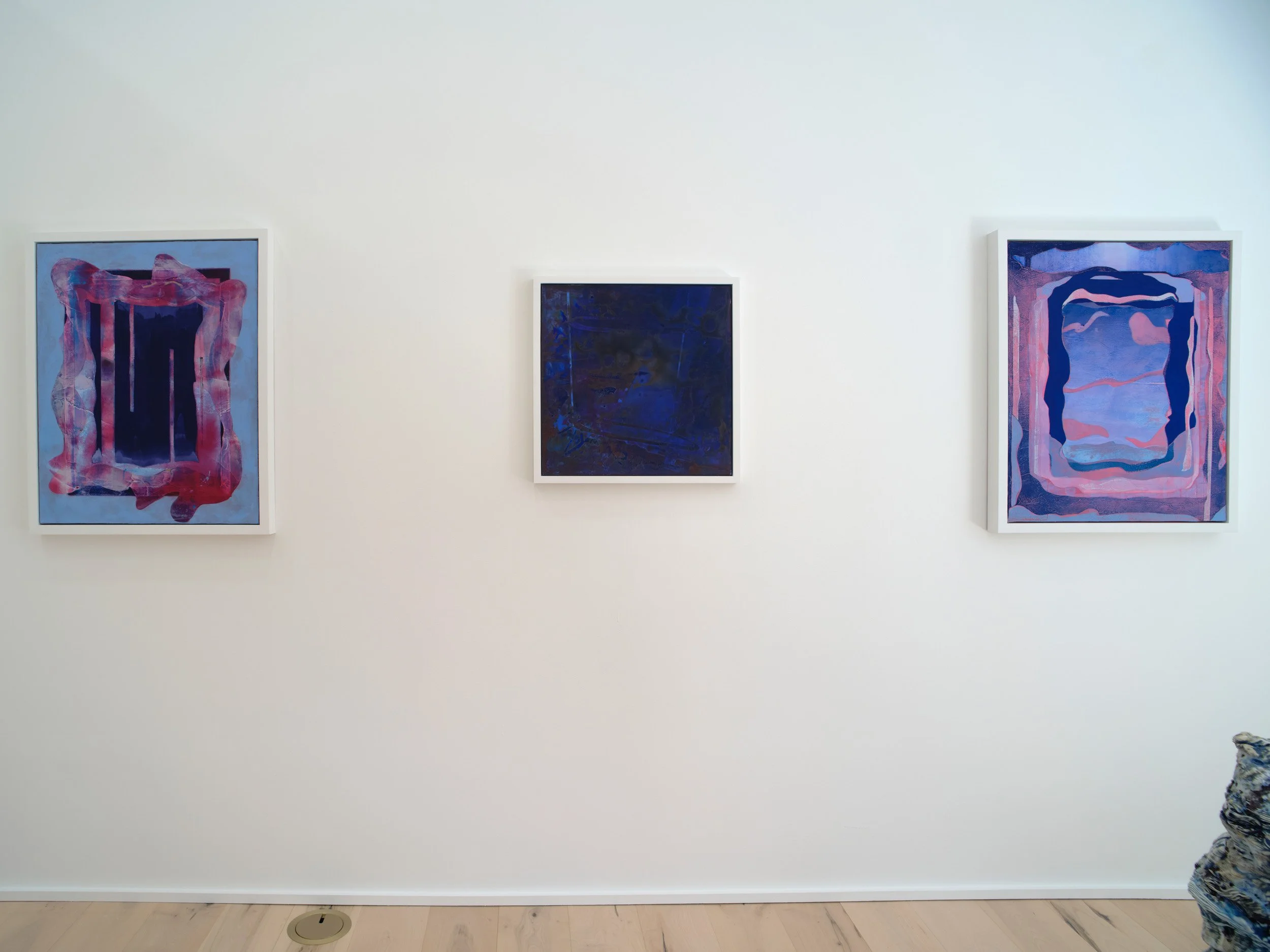 Three abstract paintings on a white wall in an art gallery. The paintings feature dark and vibrant colors with flowing, layered shapes.