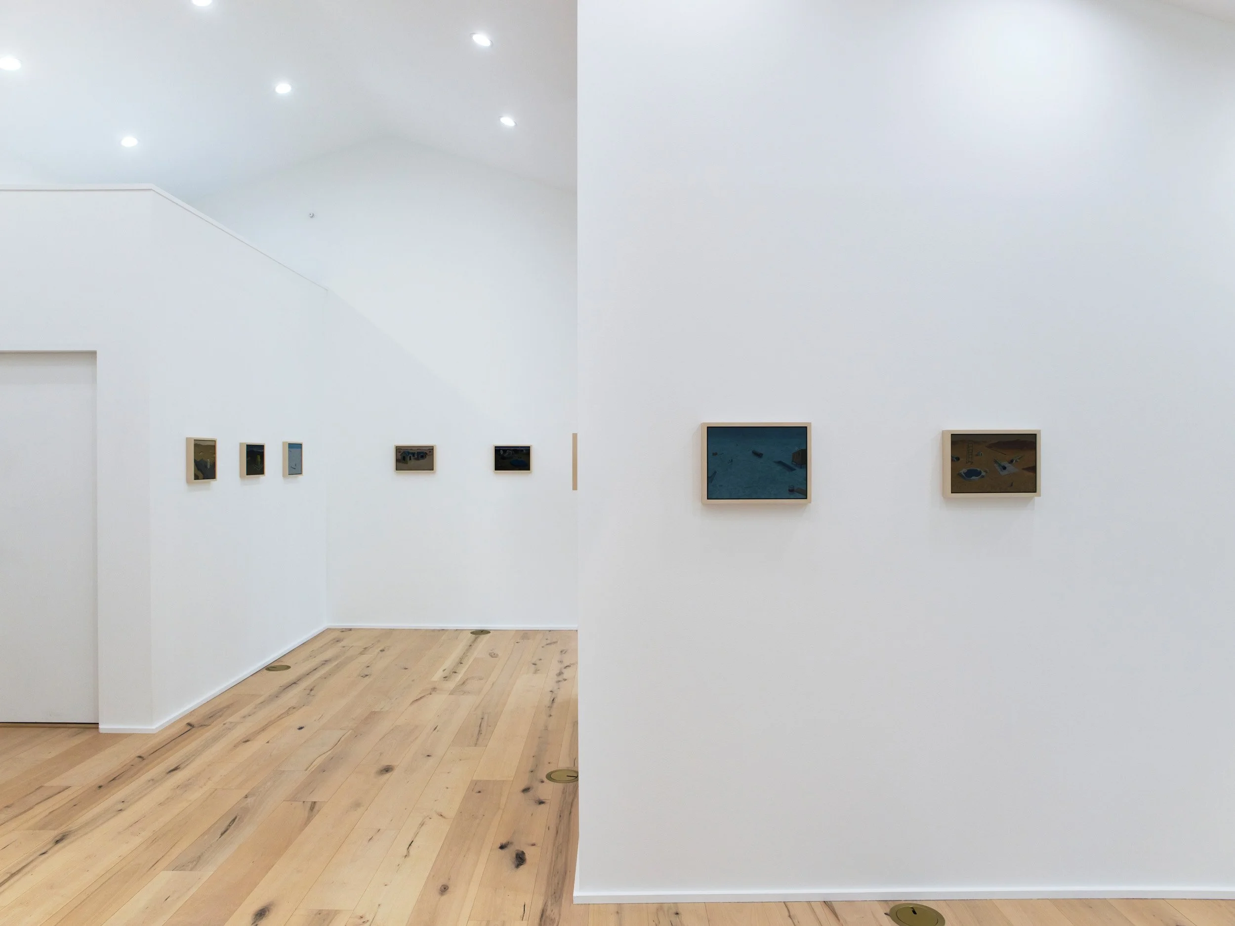 An art gallery with white walls and framed artwork, wooden floor, and bright overhead lighting.