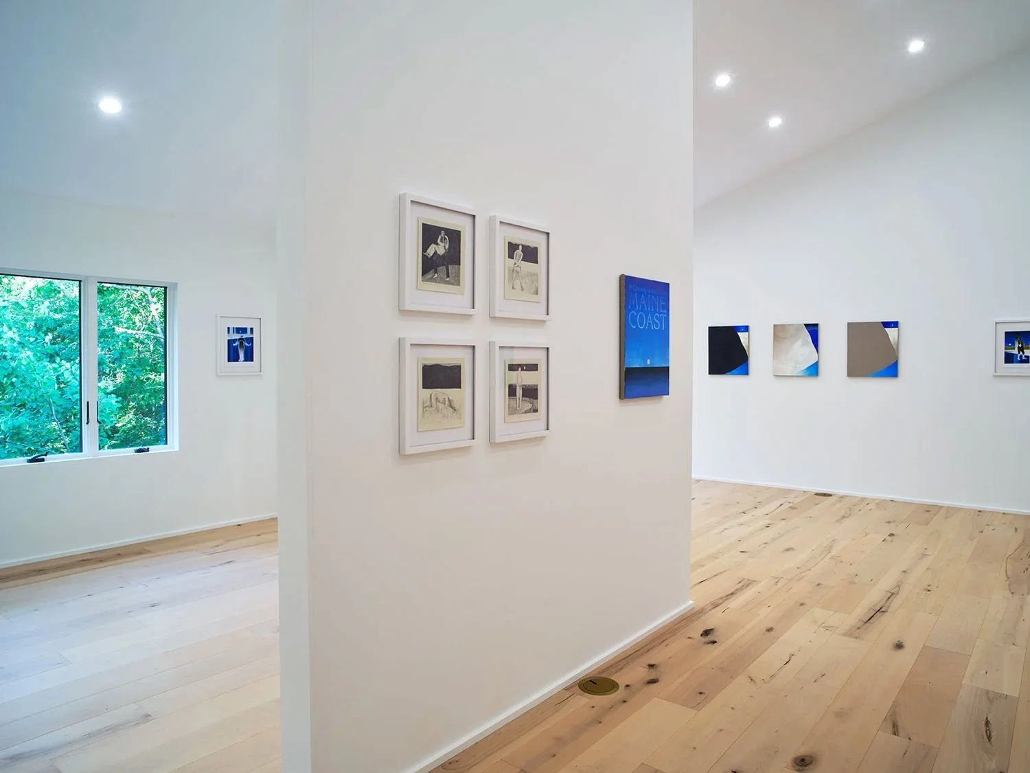 An art gallery with white walls displaying photographs and paintings, a window showing green trees outside, and wooden floors.