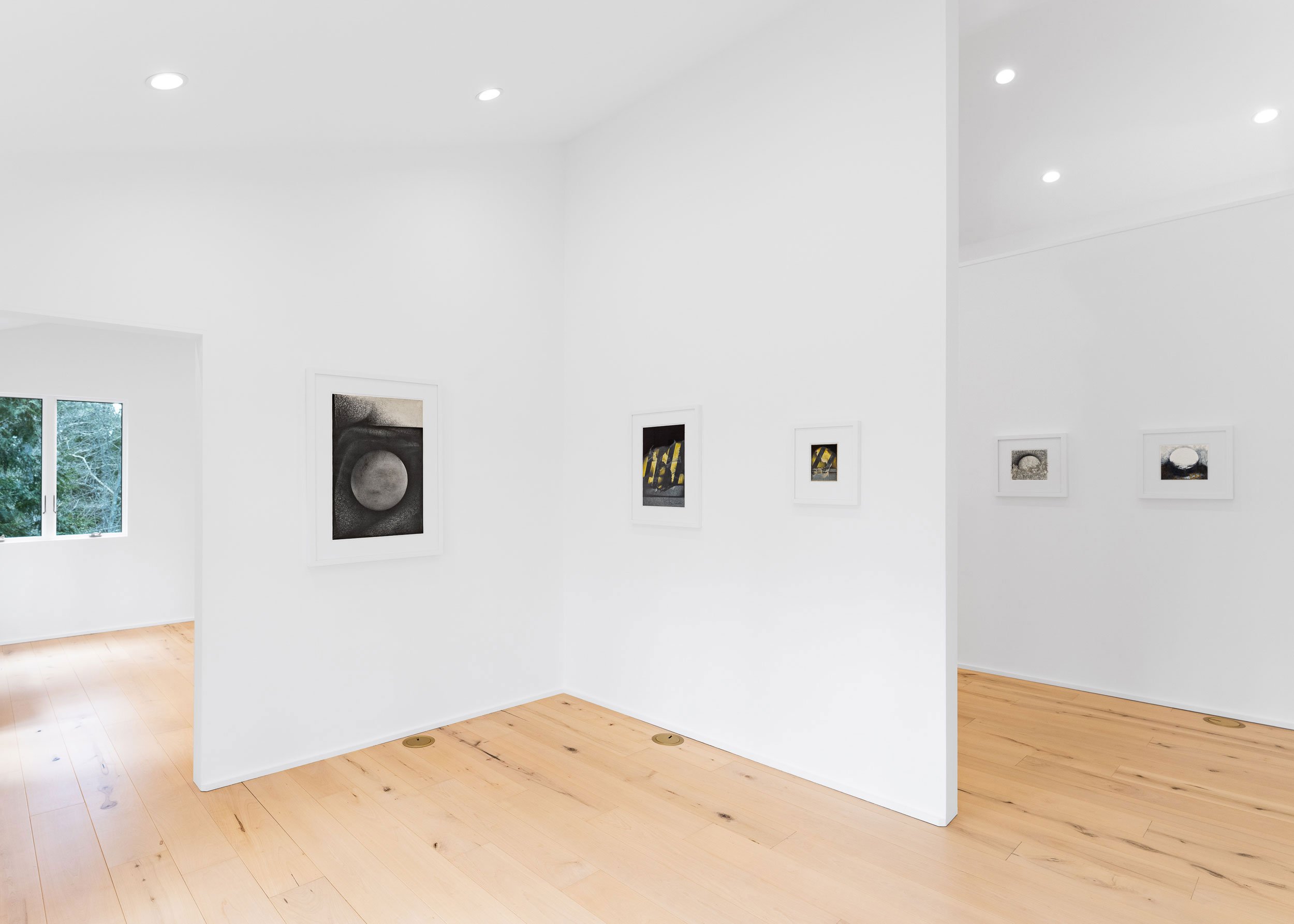 White walls in an art gallery with framed artwork, hardwood floors, and a large window with trees outside.