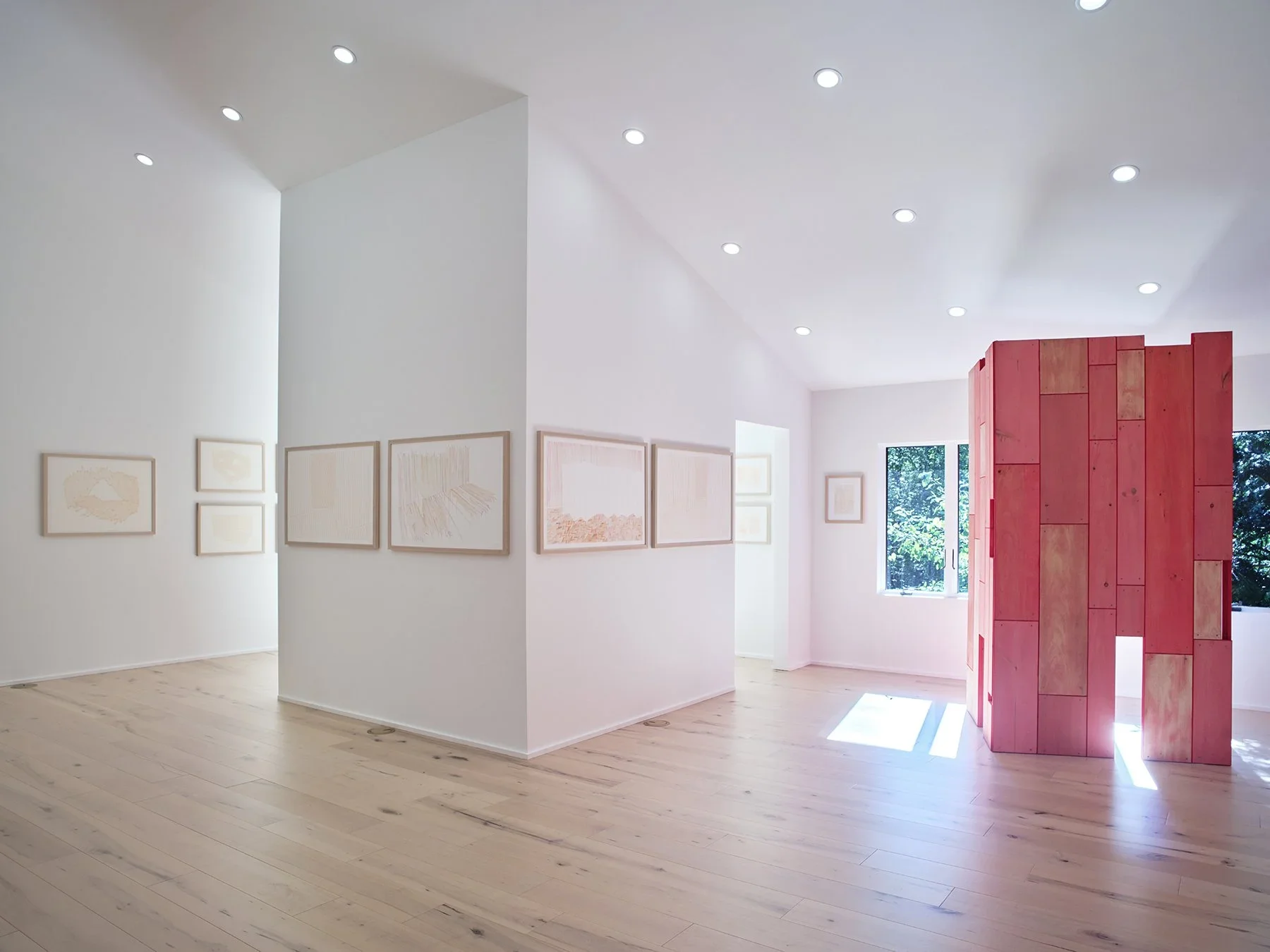 Art gallery with white walls displaying framed artwork and a pink wooden structure near the windows, with natural light coming in.