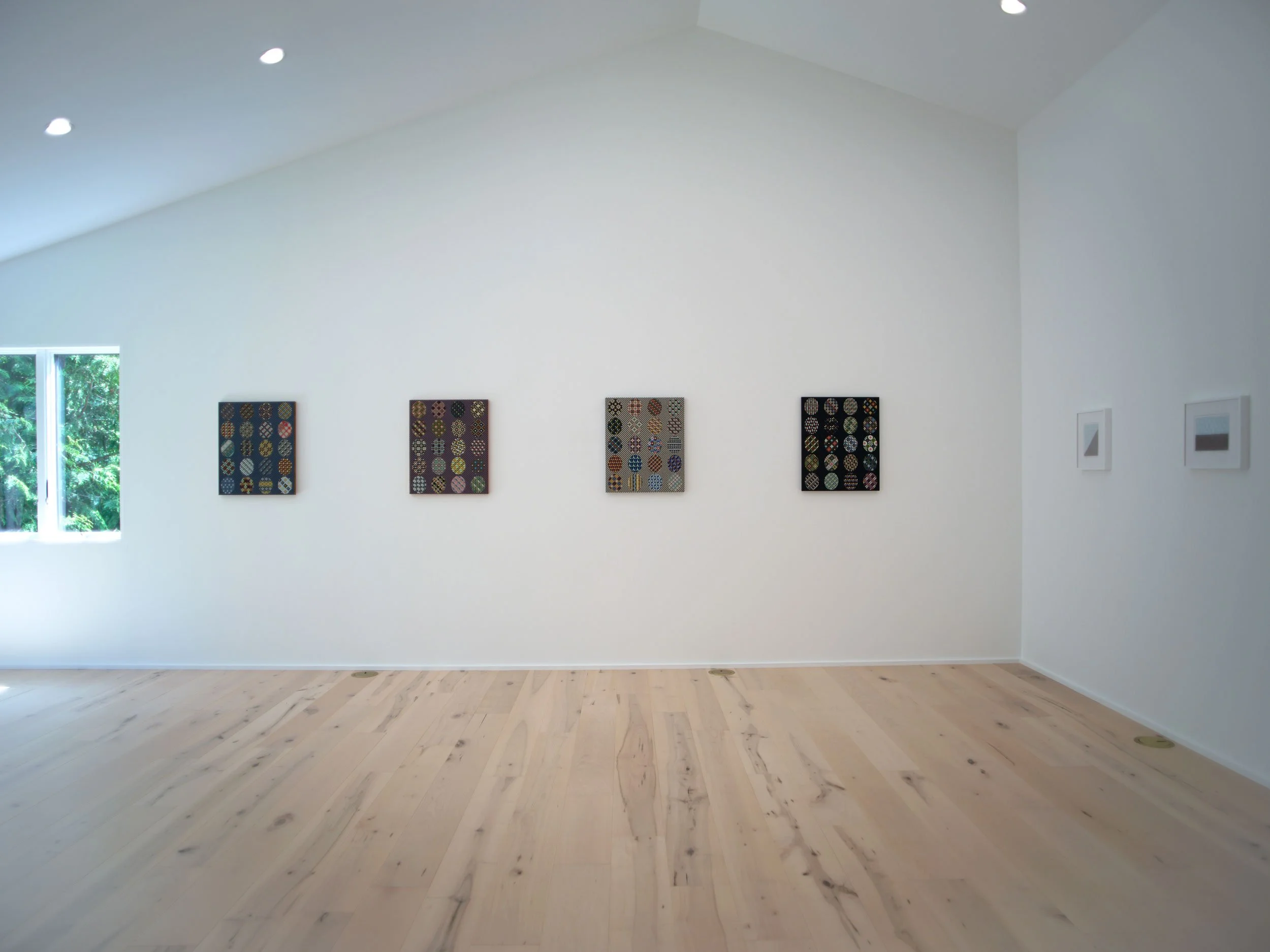 A white art gallery wall with four colorful, patterned artworks and framed photographs, and a window showing green foliage outside.