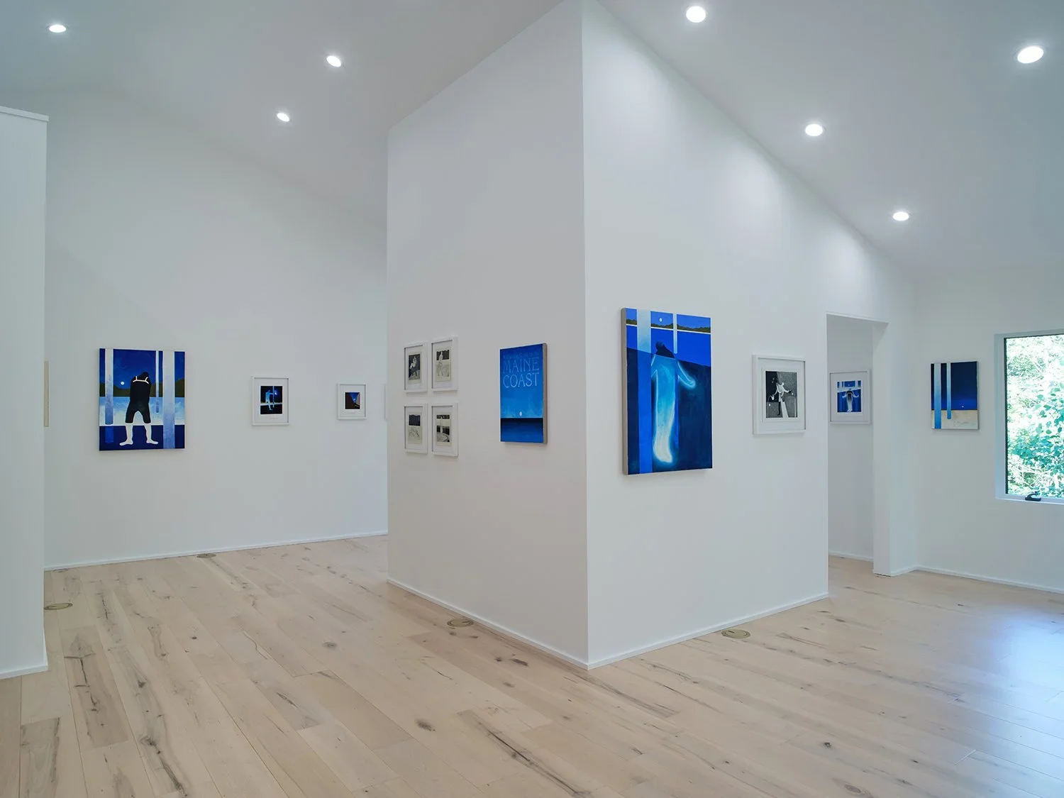 An art gallery with white walls and wooden floors displaying various framed artworks, some in black and white and others in color, with a window letting in natural light.