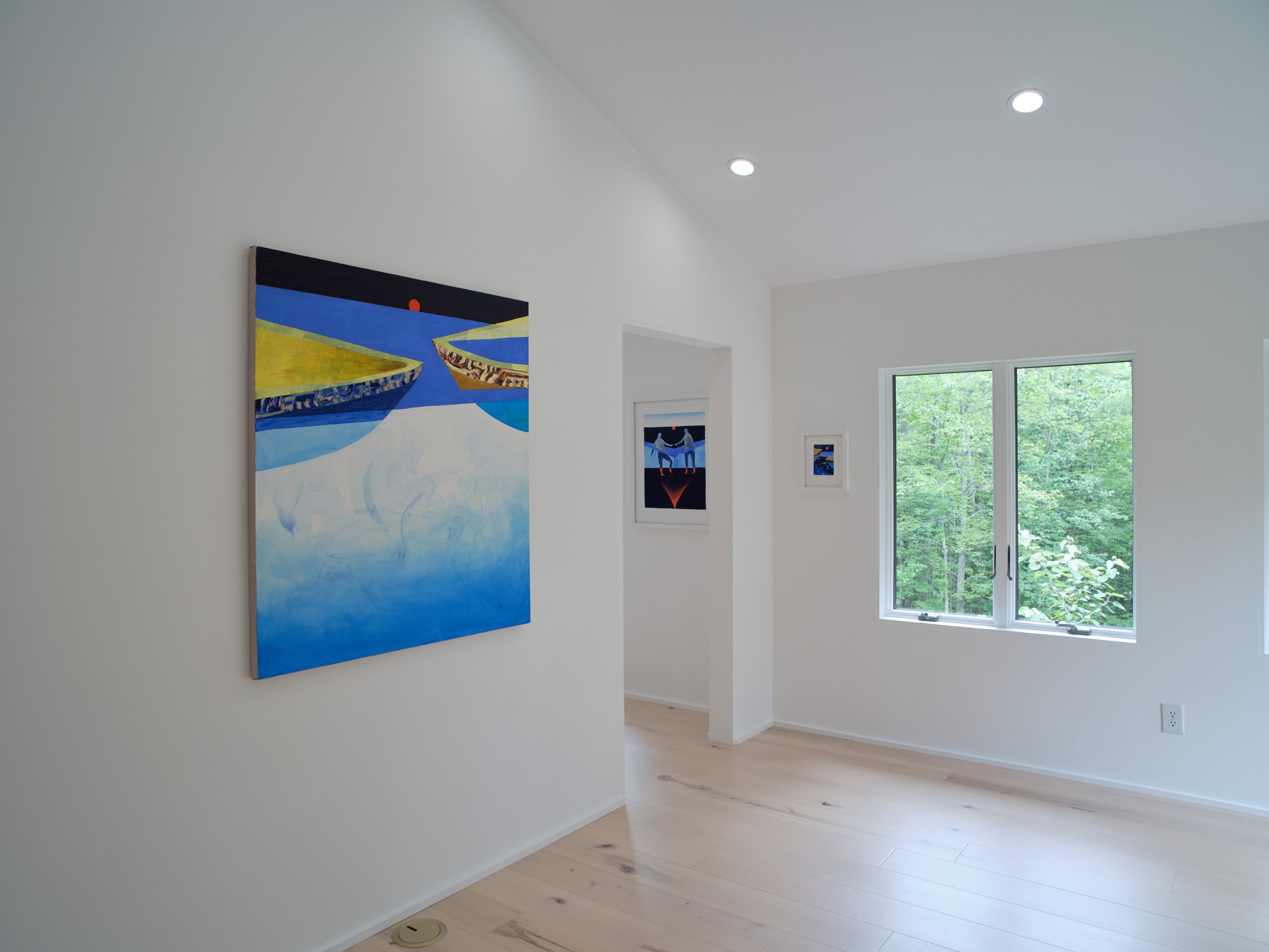 A modern white interior room with framed artwork on the walls, a large window showing greenery outside, and a hardwood floor.