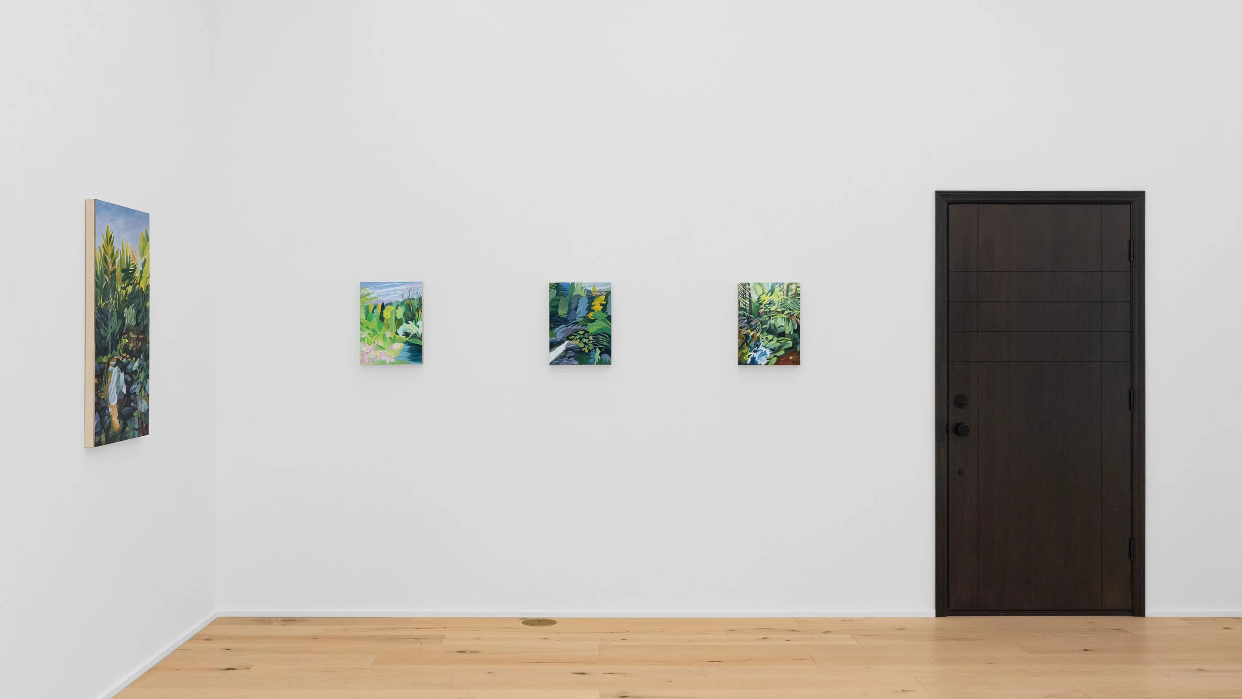 Gallery wall with four colorful landscape paintings in an art gallery with a white wall, black door, and wooden floor.