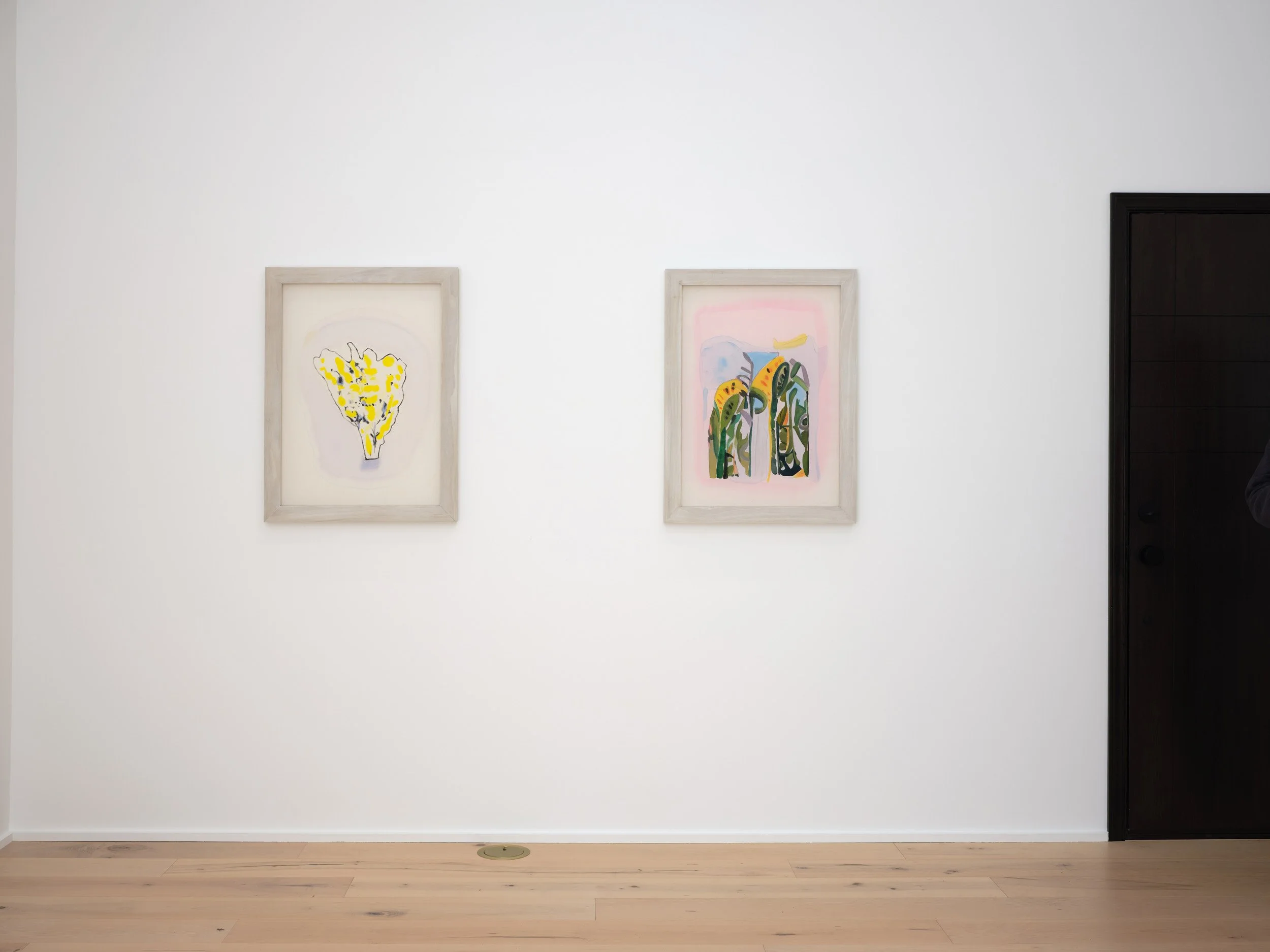 Two framed abstract paintings hang on a white gallery wall, with a black door to the right. The paintings feature colorful, organic shapes with one in yellow and black, and the other with green, yellow, and blue elements.