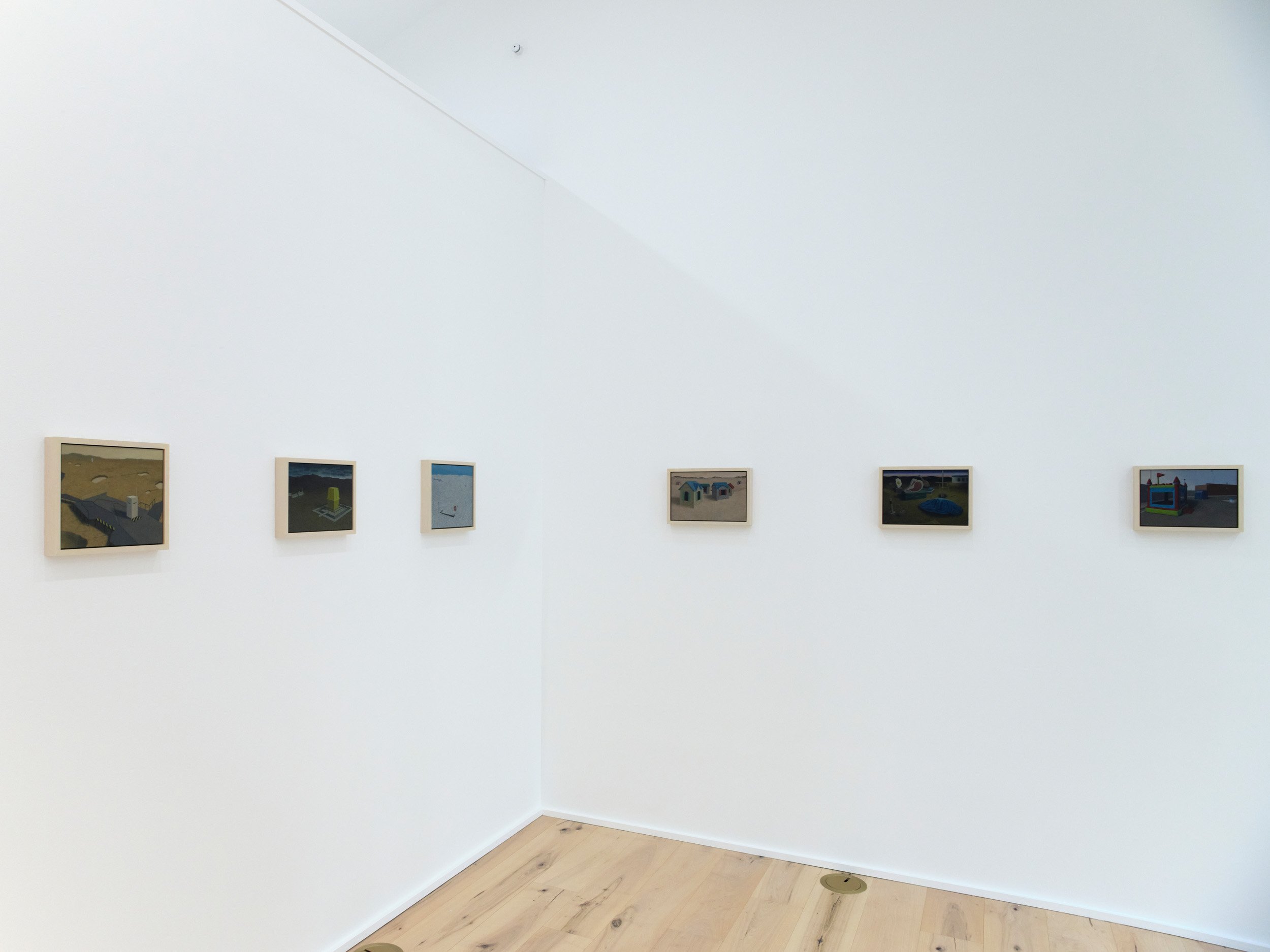An art gallery white wall with a collection of framed paintings arranged in a row at eye level, with light wooden flooring below.