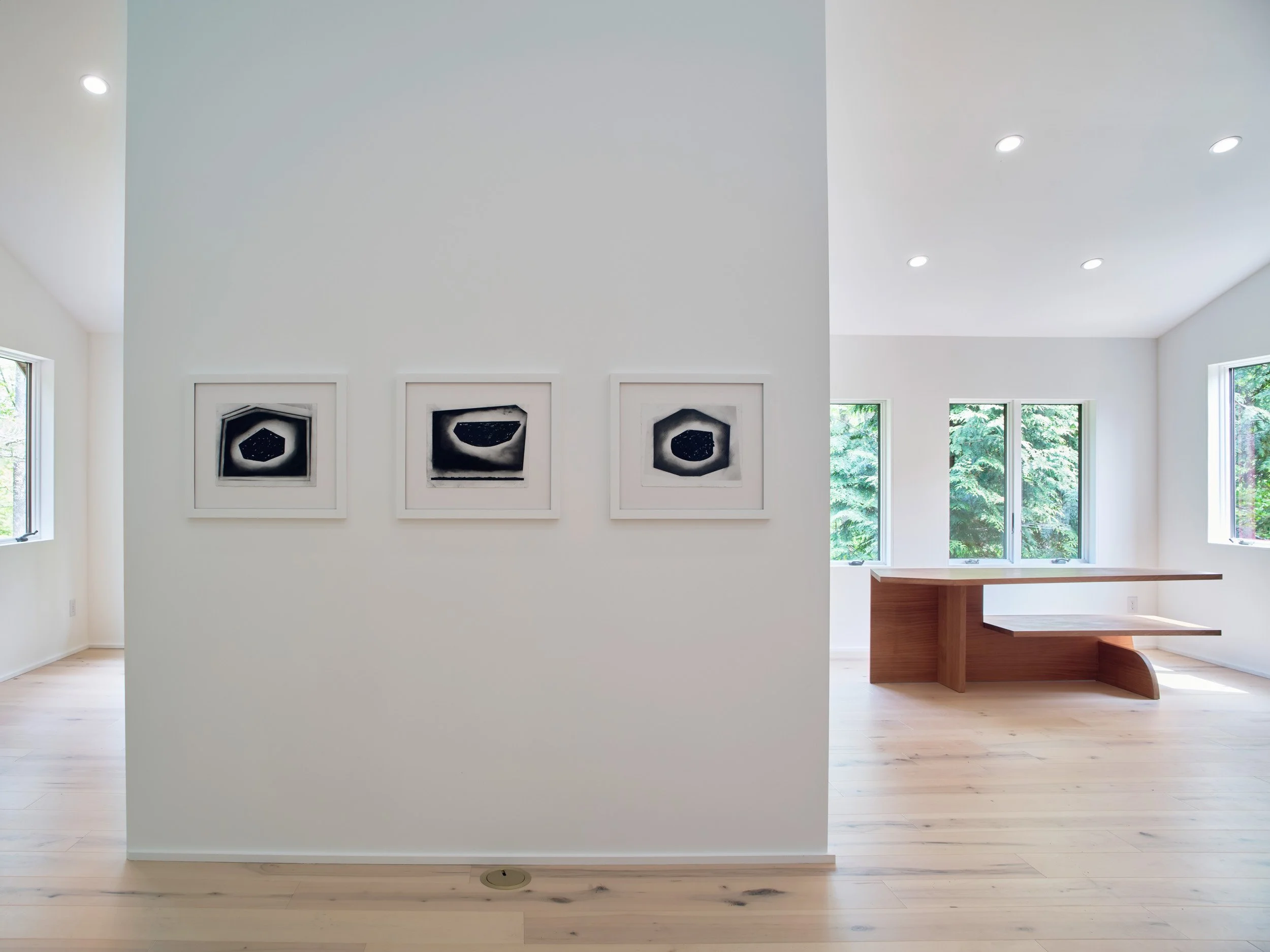 Interior view of a modern, white-walled room with three black and white framed artworks on the left wall. Large windows on the right show green foliage outside, and there is a built-in wooden bench under the windows. The ceiling has multiple recessed lights, and the floor is light-colored wood.