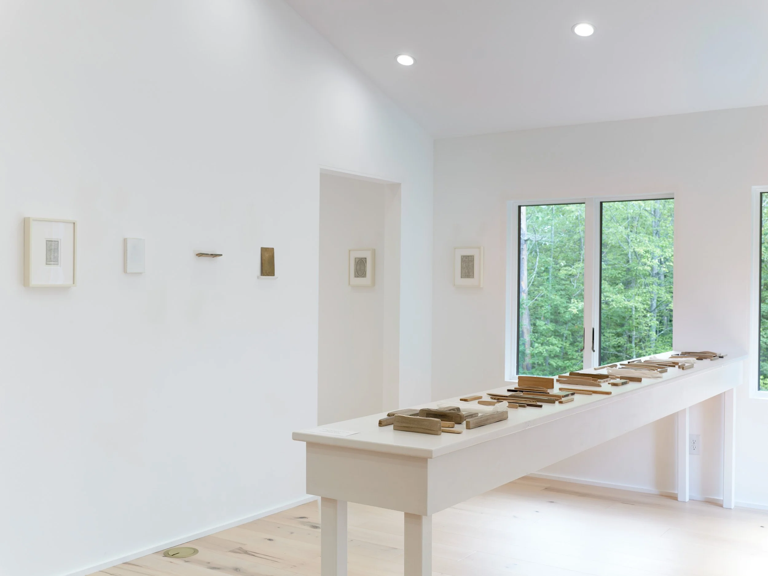 Contemporary art gallery with white walls, framed artwork, wooden sculptures on a table, and large windows showing green trees outside.
