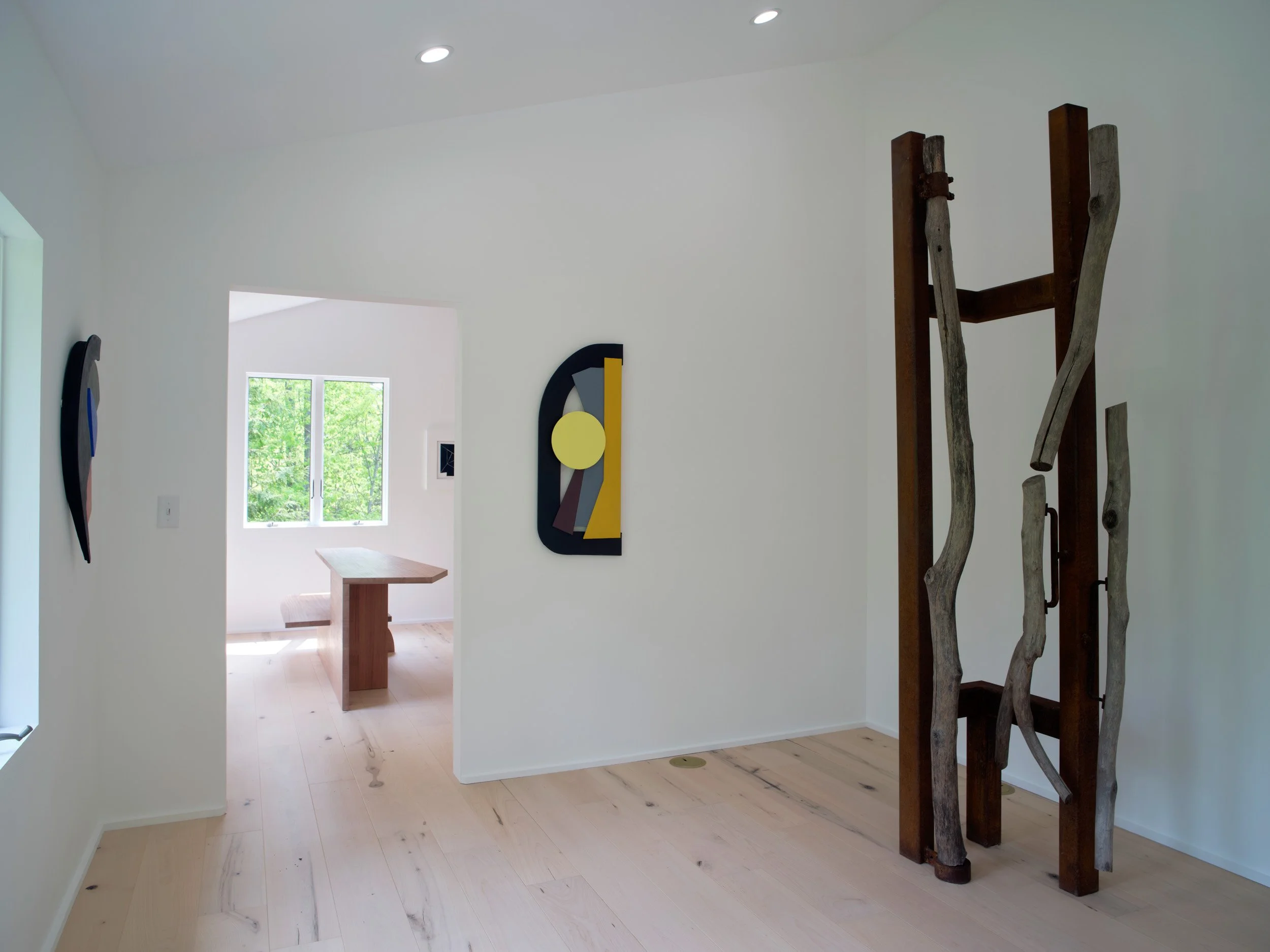 A room with white walls, light wood flooring, and a window showing green trees outside. Decor includes modern abstract art on the walls and a wooden table. There are wooden sculptures or driftwood pieces on a dark wooden frame on the right side.