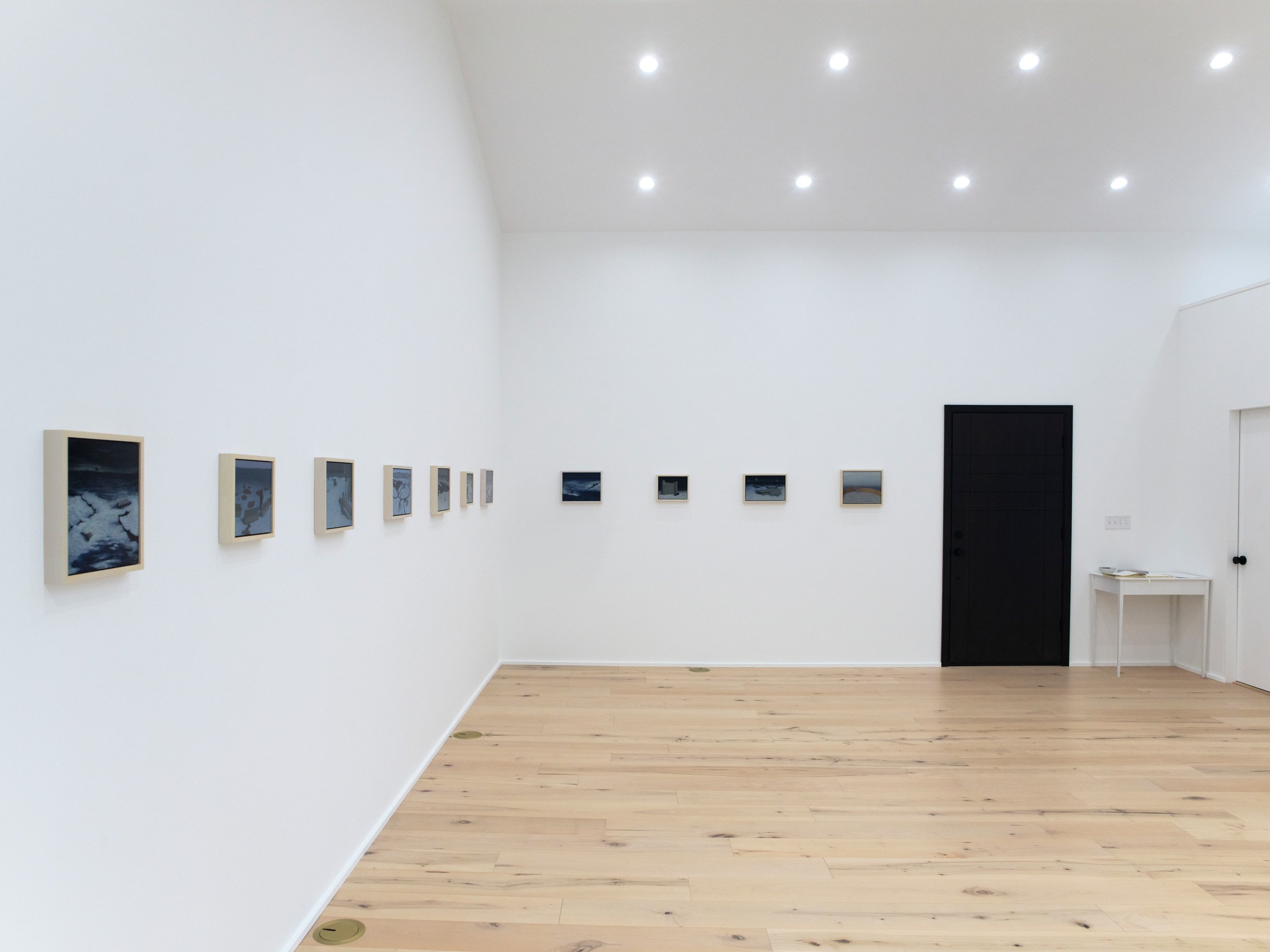 A gallery room with framed artwork hanging on white walls, a wooden floor, and a black door.