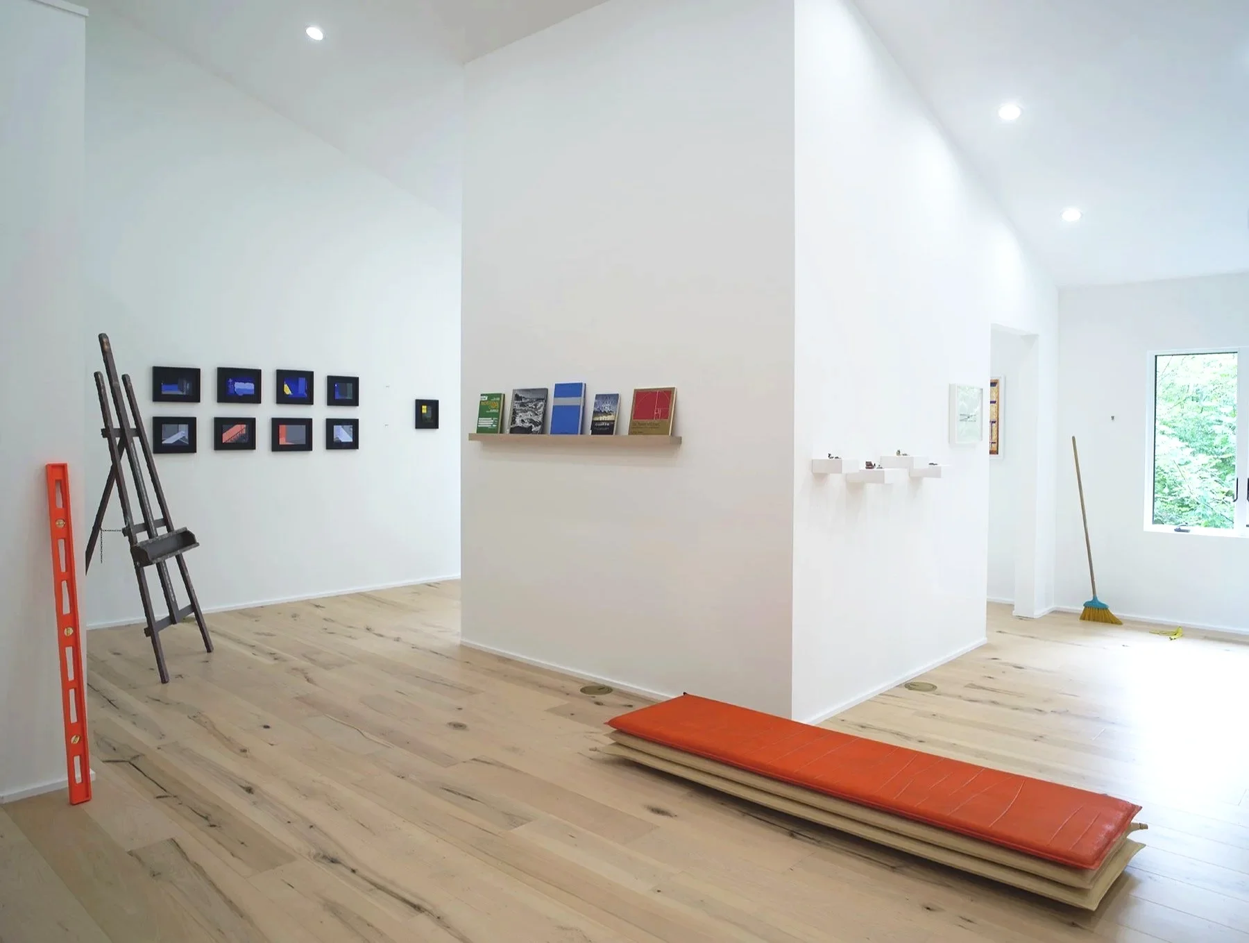 Interior of a modern, minimalistic house with white walls and light wooden flooring. Construction or renovation tools and materials, including a ladder, a broom, an orange cushion, and framed artwork, are visible. A floating shelf with books and a window showing green trees are also present.