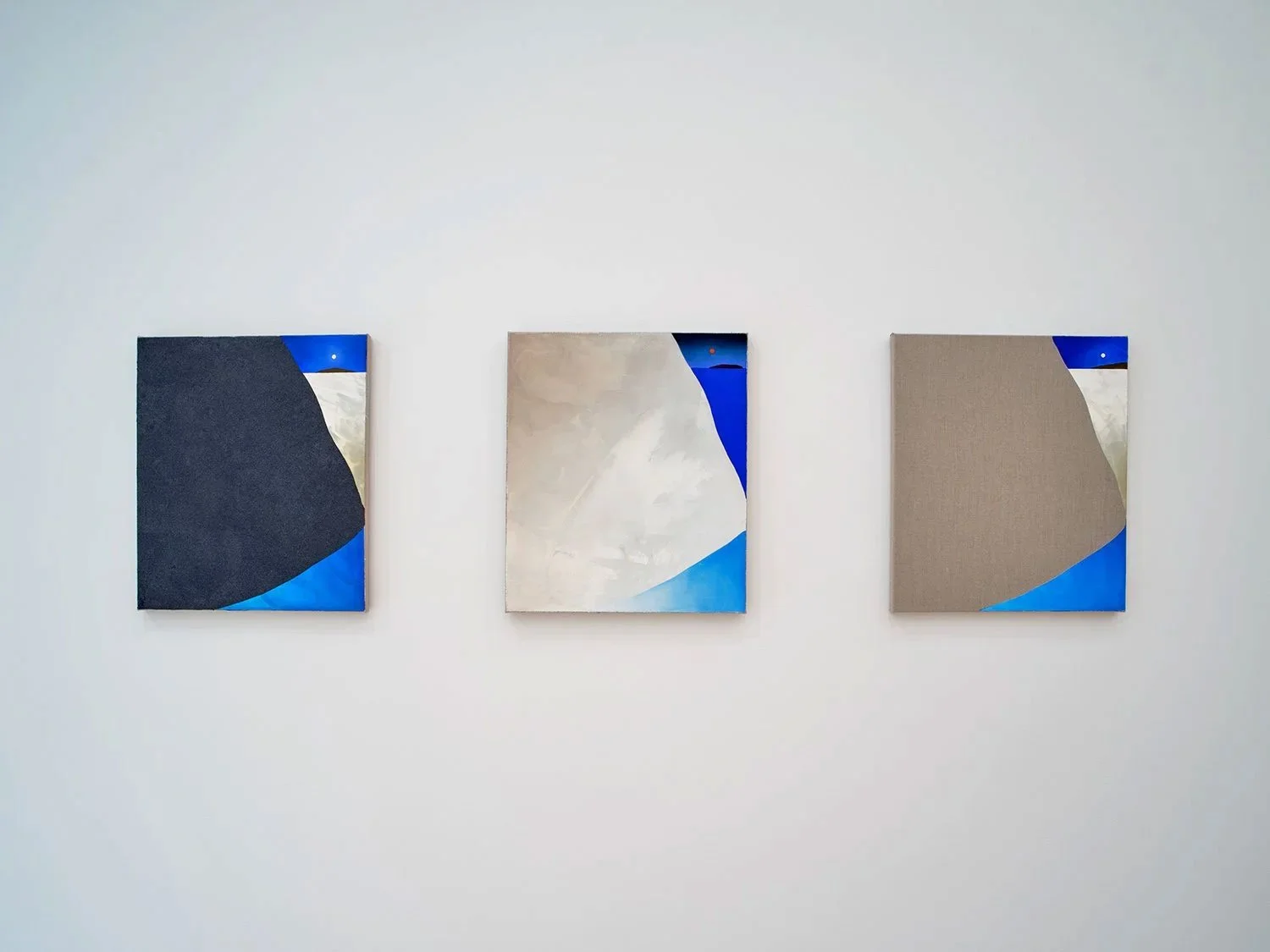 Three geometric abstract paintings hanging on a white gallery wall, each with shades of gray, blue, black, and beige.
