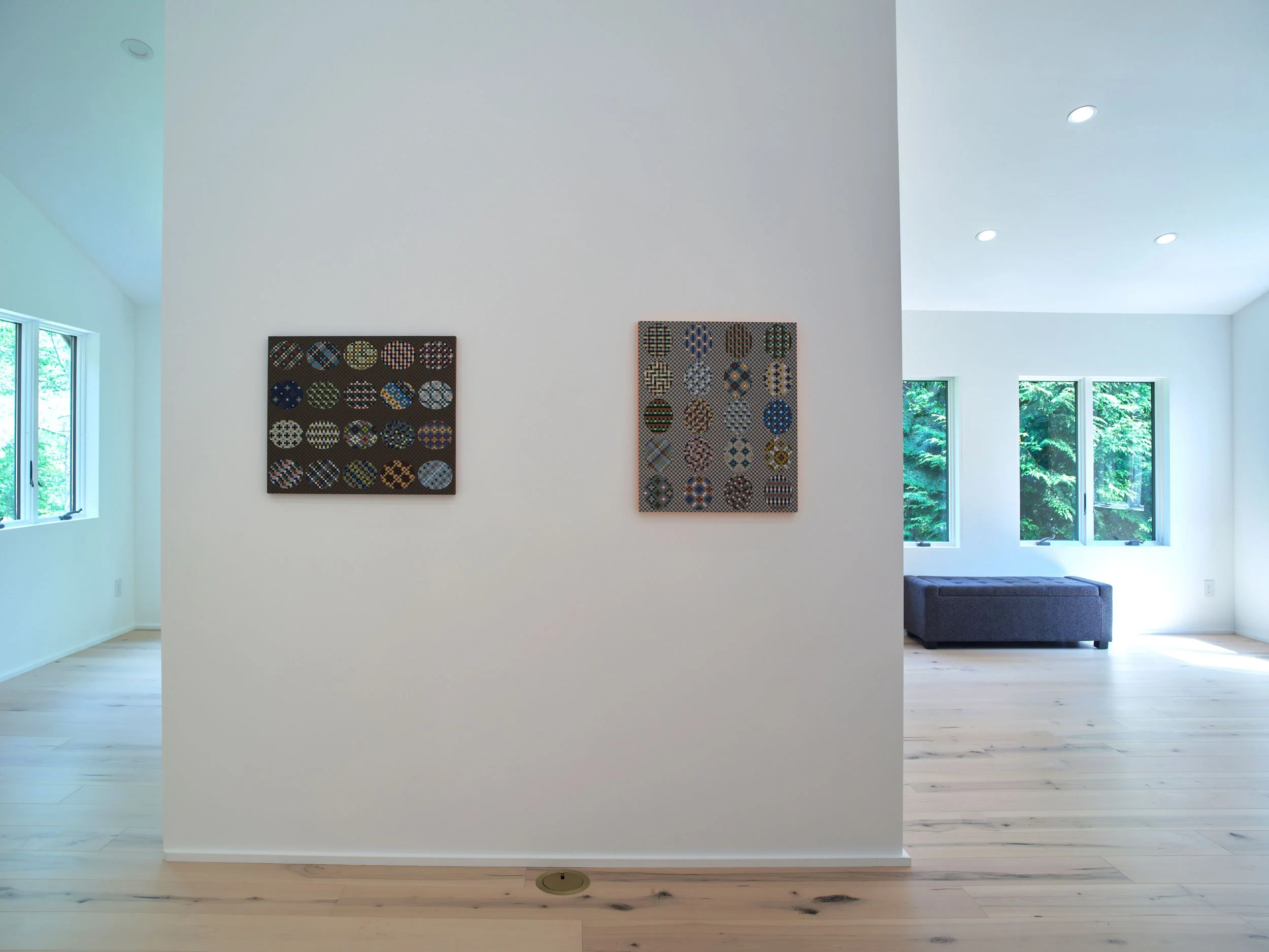 A minimalistic art gallery with two colorful patterned artworks hanging on a white wall, large windows allowing natural light, and light wood flooring.