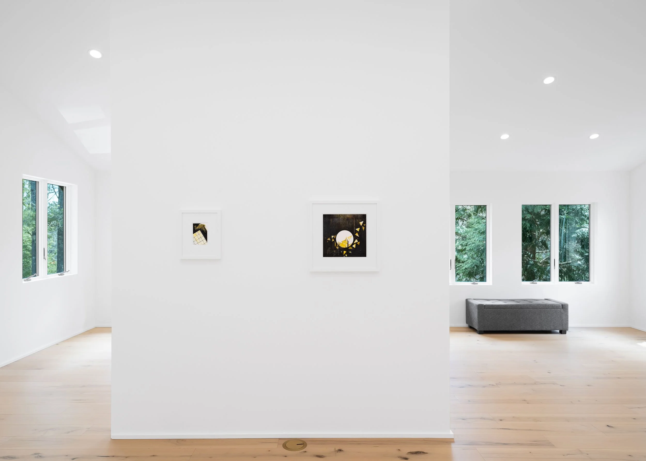 Modern white art gallery with artworks on walls, large windows showing green trees outside, light wood flooring, and a gray sofa.