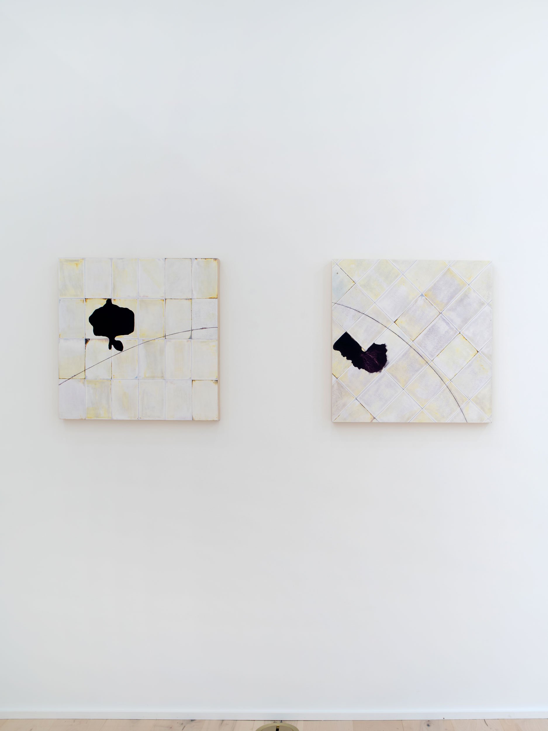 Two abstract paintings with black shapes on a white tiled background are hung on a white wall.
