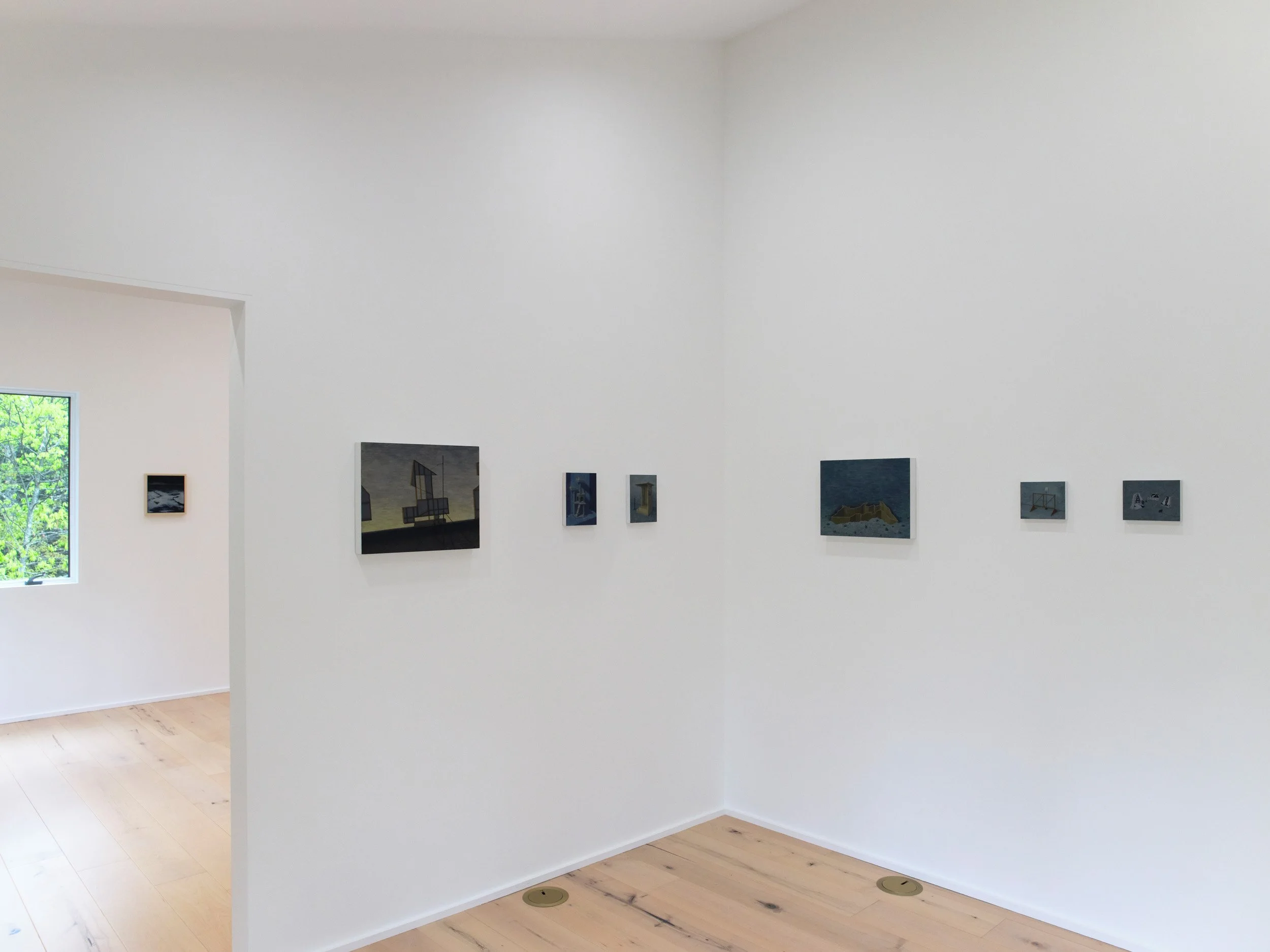 An art gallery with various paintings hanging on white walls and a wooden floor, with a window showing green trees outside.