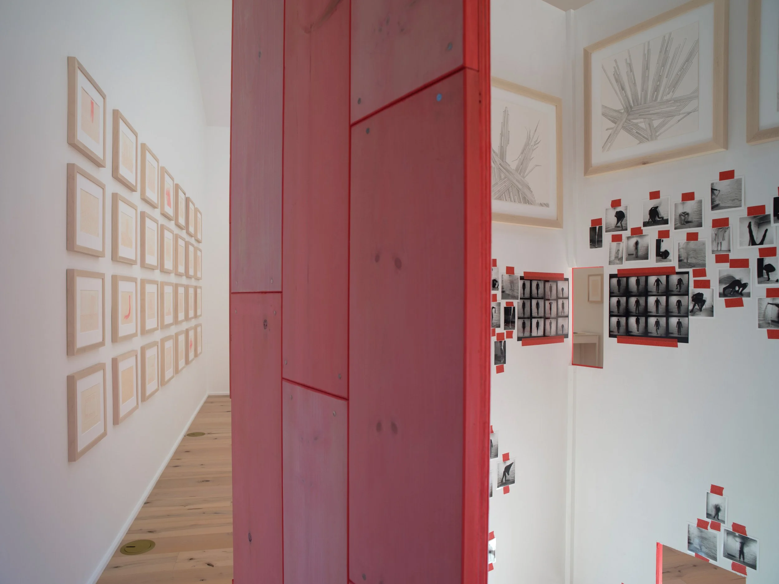 An art gallery with framed pictures on white walls and black and white photographs displayed with red tape. A large red wooden structure is positioned in the middle of the room, partially obstructing the view.