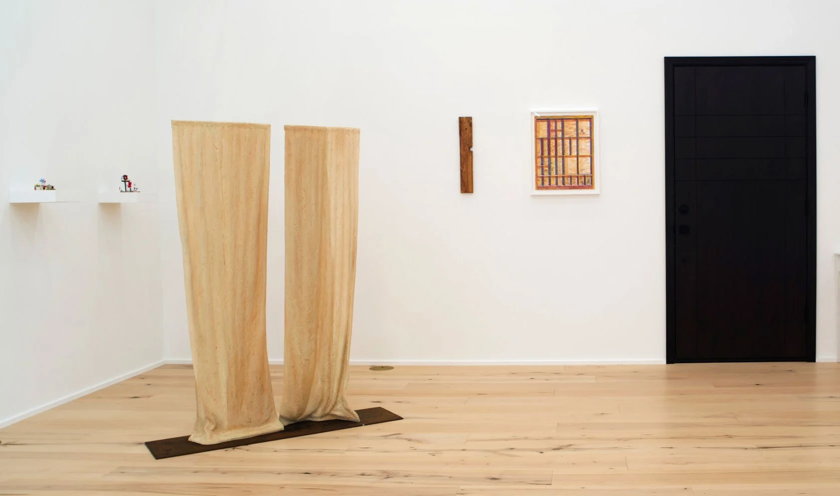 Art gallery with two wooden sculptures, small framed artwork, a vertical wooden piece on the wall, and a black door.