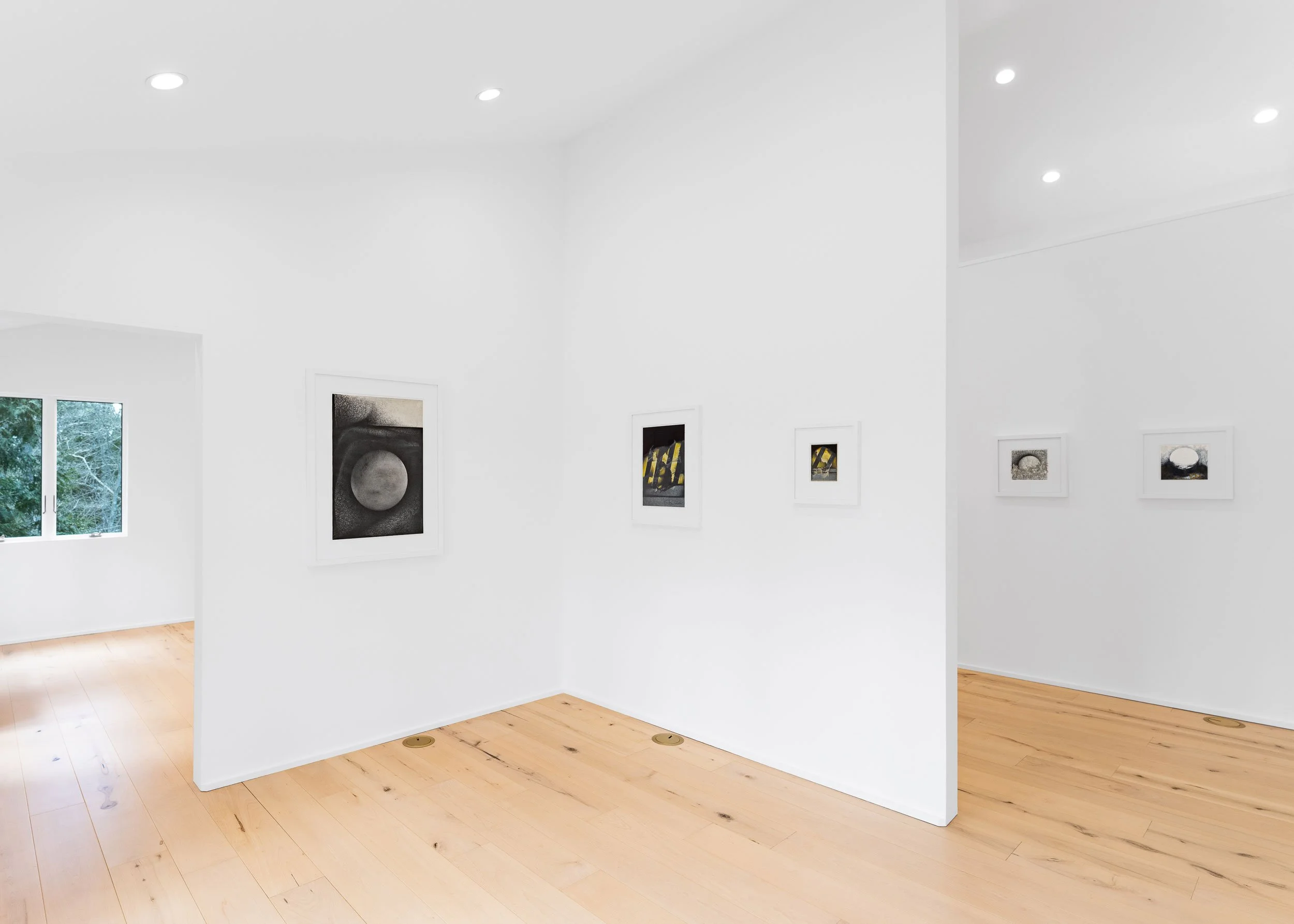 A modern art gallery with white walls, wooden floors, and four pieces of framed artwork hanging on the walls. A window shows green trees outside.
