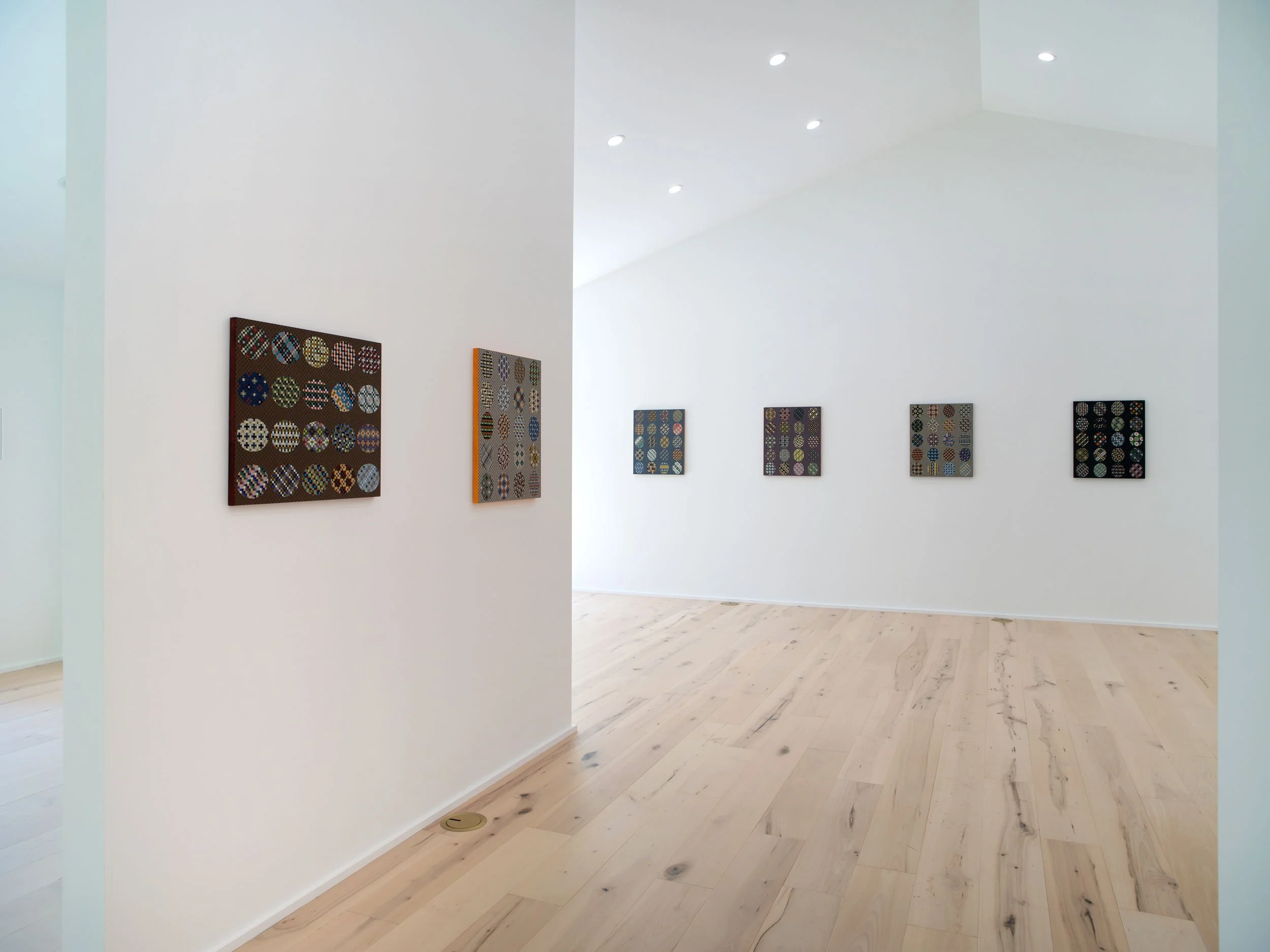 A gallery with white walls displaying multiple colorful geometric patterned art pieces, hardwood floors, and ceiling spotlights.