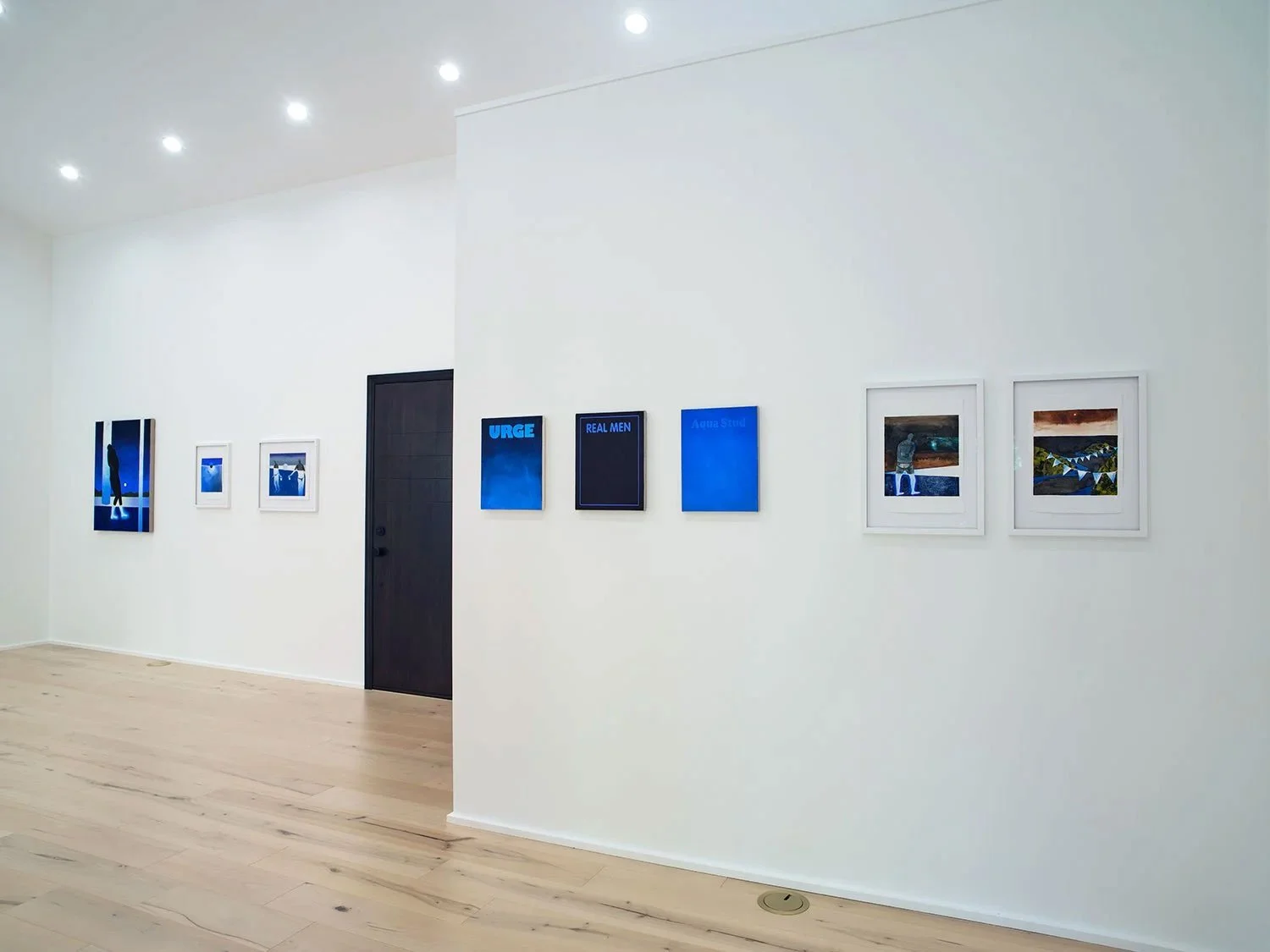 Gallery wall with nine framed art pieces and digital screens on a white wall, with light wood flooring and ceiling lights.