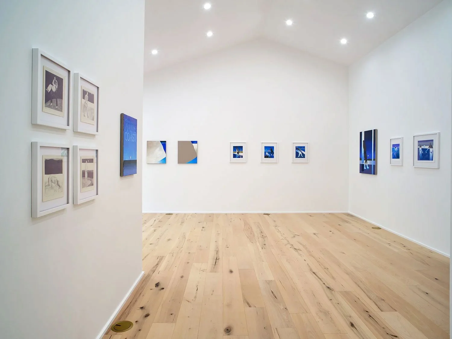 Interior of an art gallery with white walls, light wood flooring, and framed abstract artworks in shades of blue and grey displayed throughout, illuminated by ceiling lights.