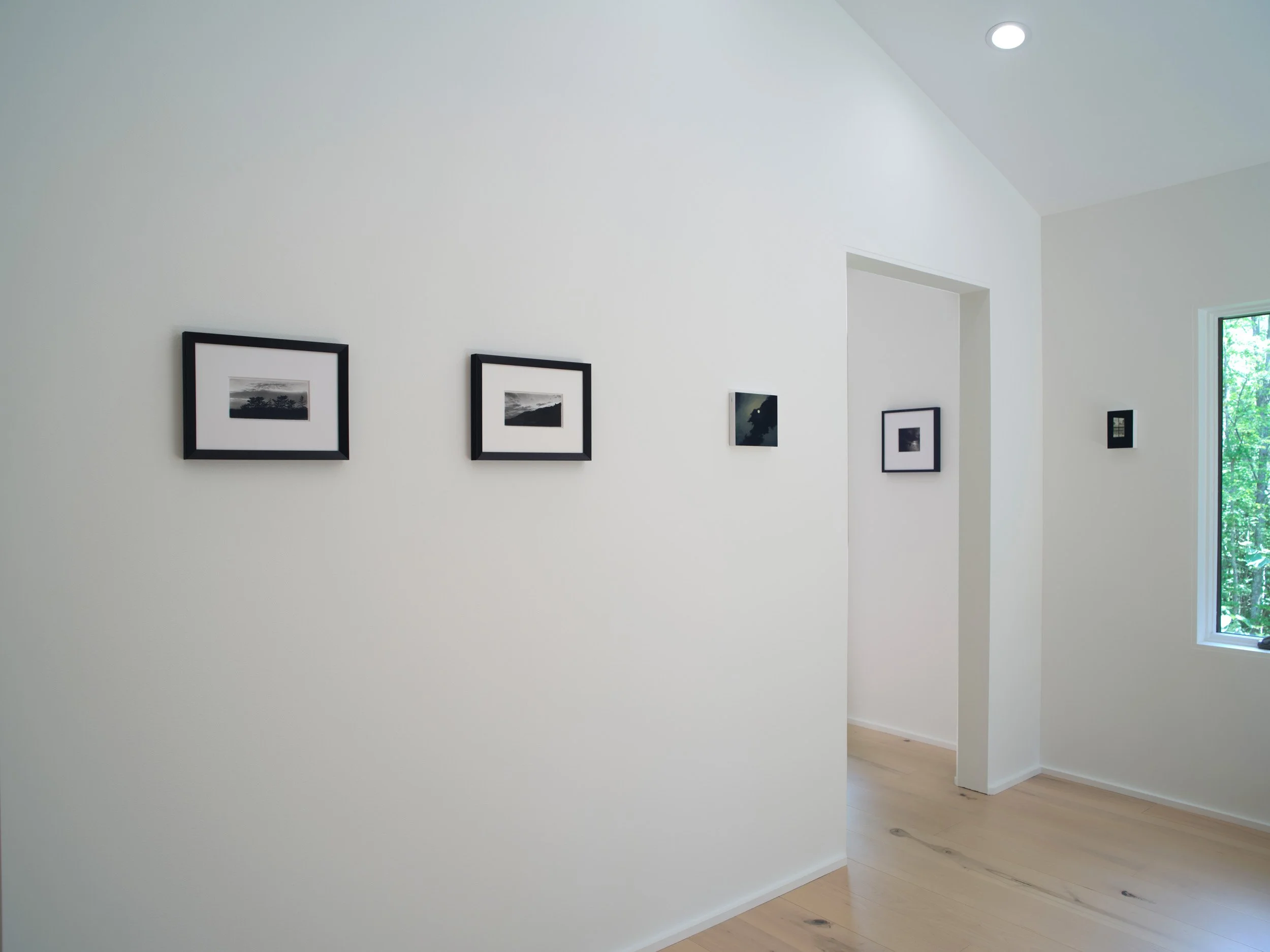 Interior of a modern home with white walls, framed black and white photographs, a large window showing greenery outside, and wooden flooring.